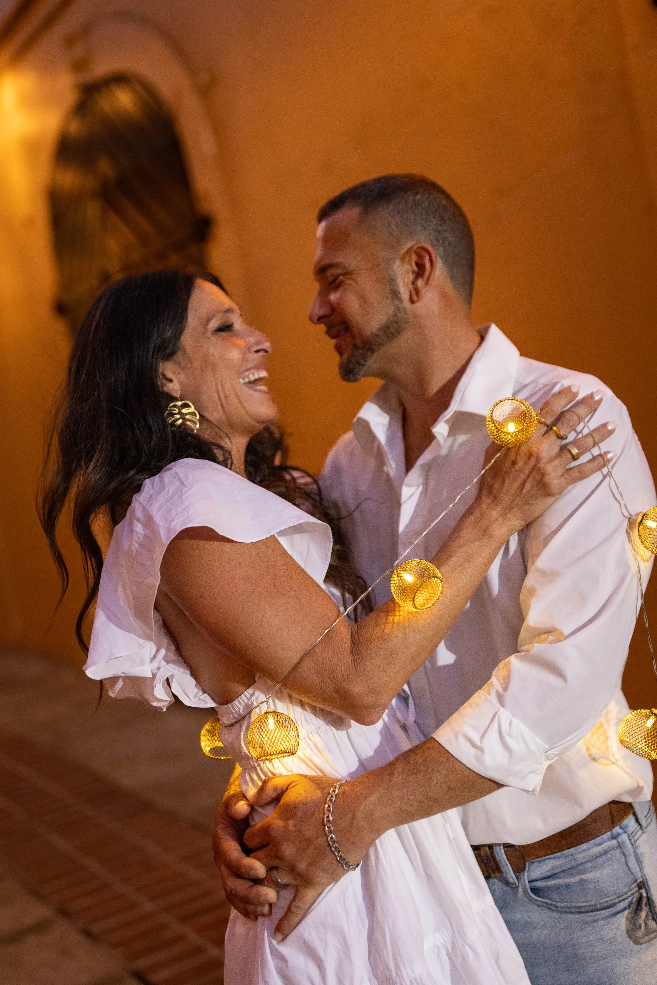 A couple dancing together, smiling and looking at each other, surrounded by warm lighting and decorative string lights.