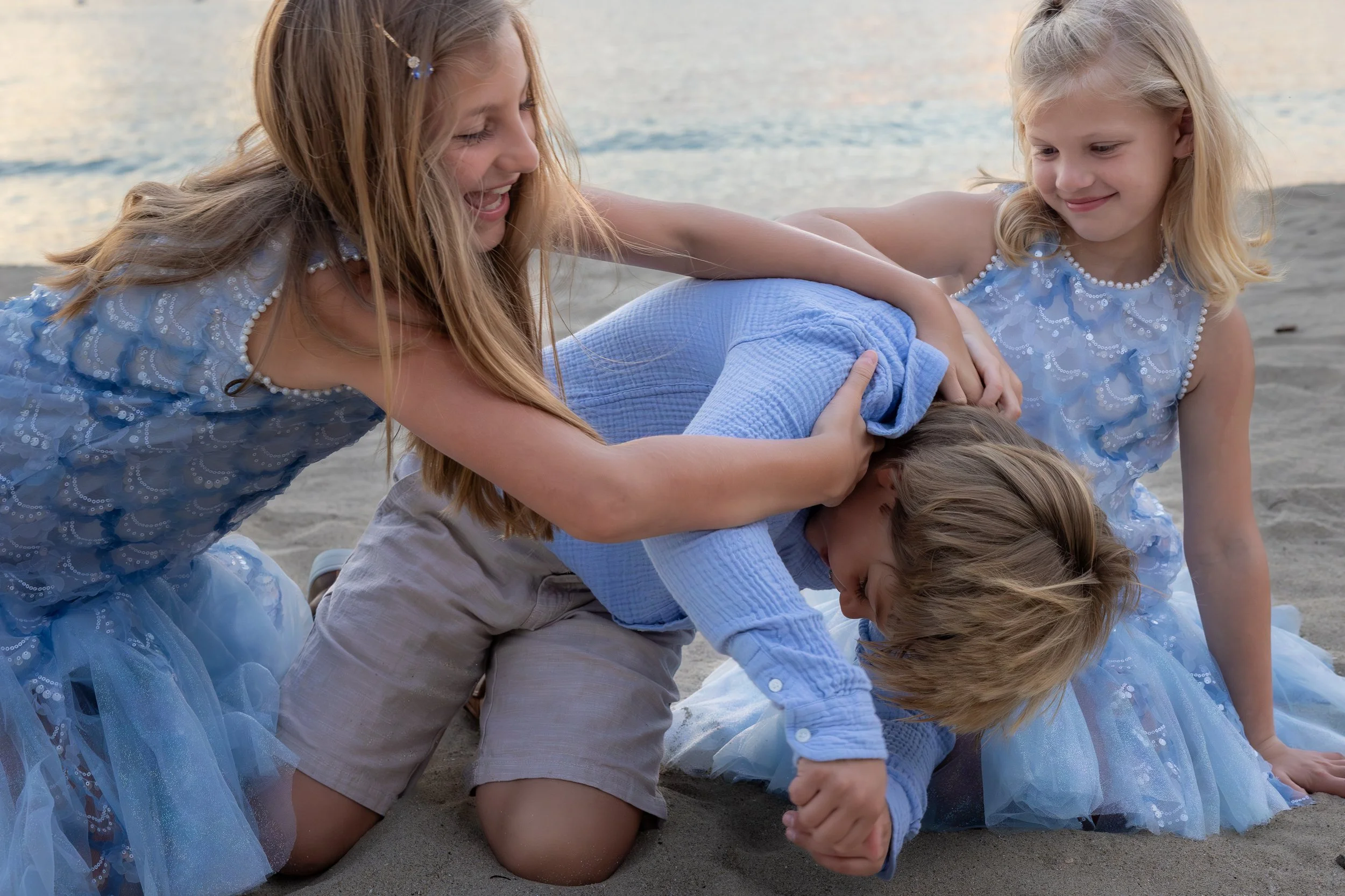 Four kids, two girls and two boys, dressed in blue clothes, playfight on a sandy beach near the water during sunset.