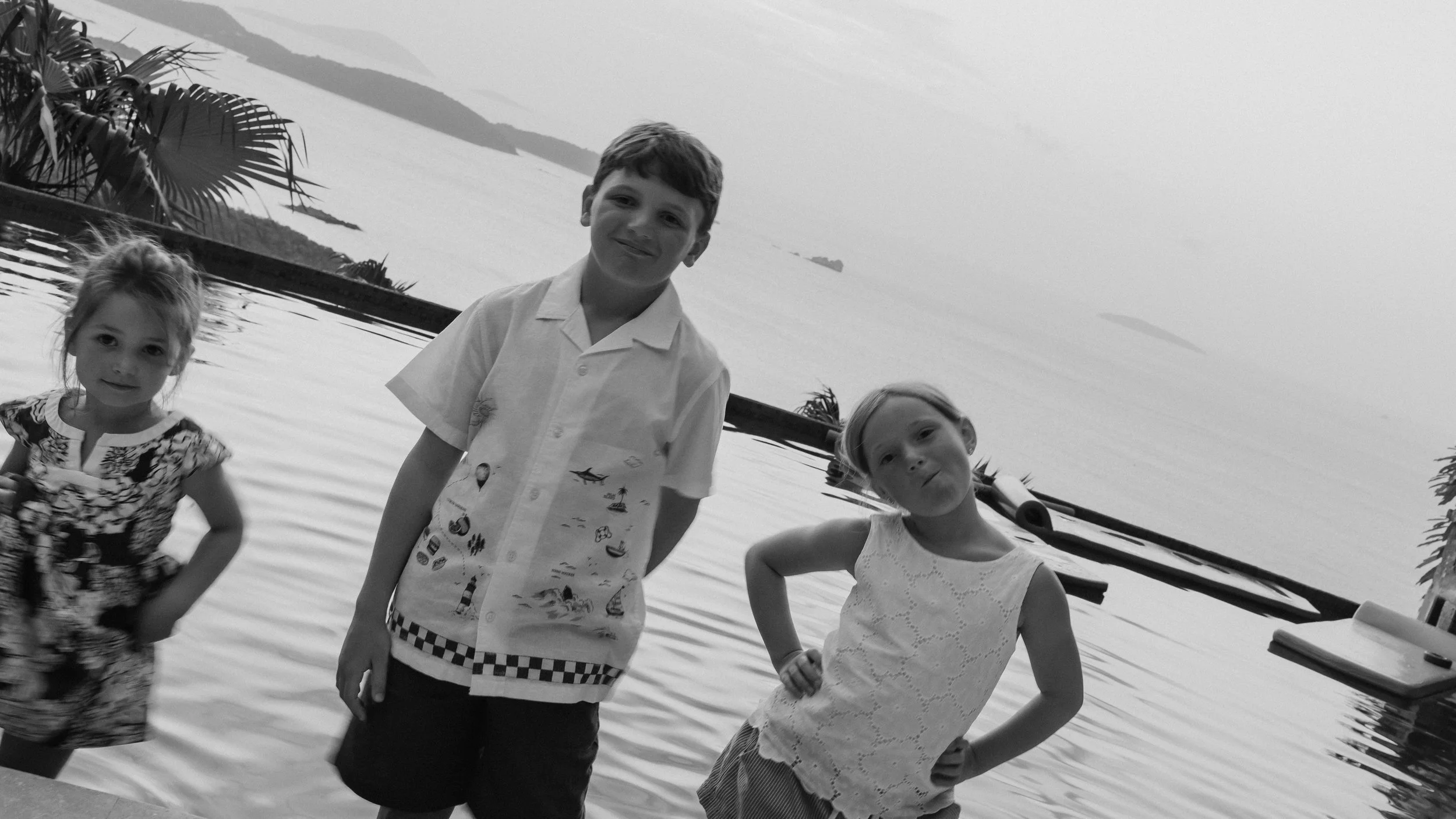 Three children standing by the water with a boat in the background, overlooking a lake and distant islands, in black and white.