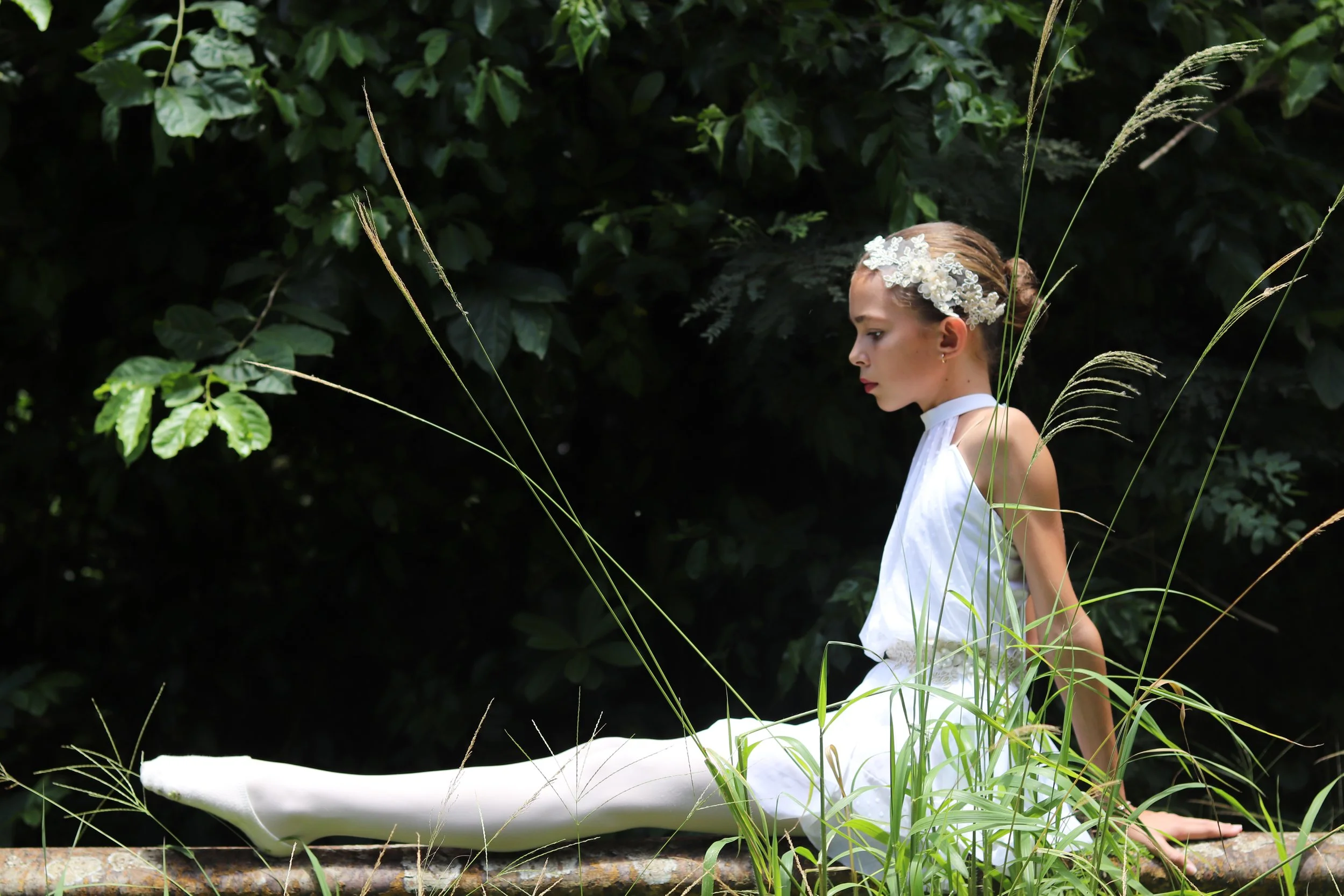 Young woman in a white dress with floral headpiece sitting on a log in a forest with tall grass and green foliage surrounding her.