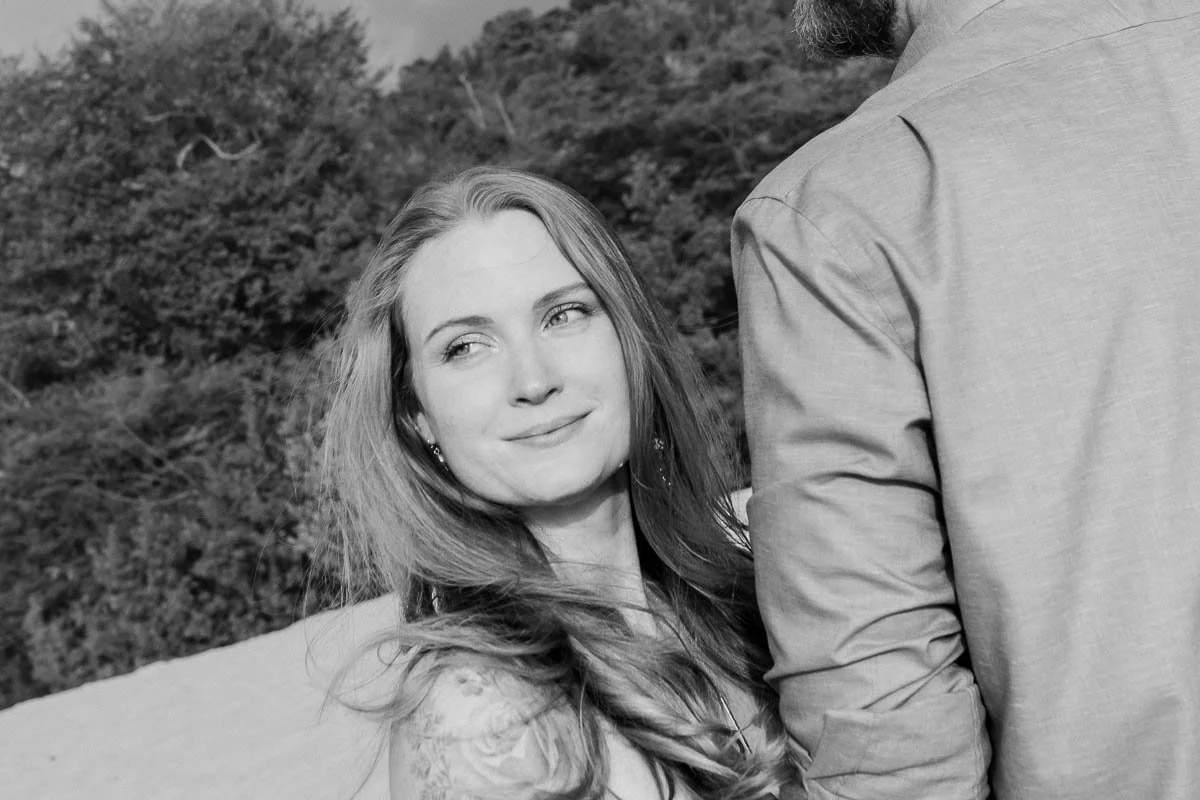 A woman looking at a man, smiling softly. They are outdoors with trees and shrubs in the background. The photo is in black and white.