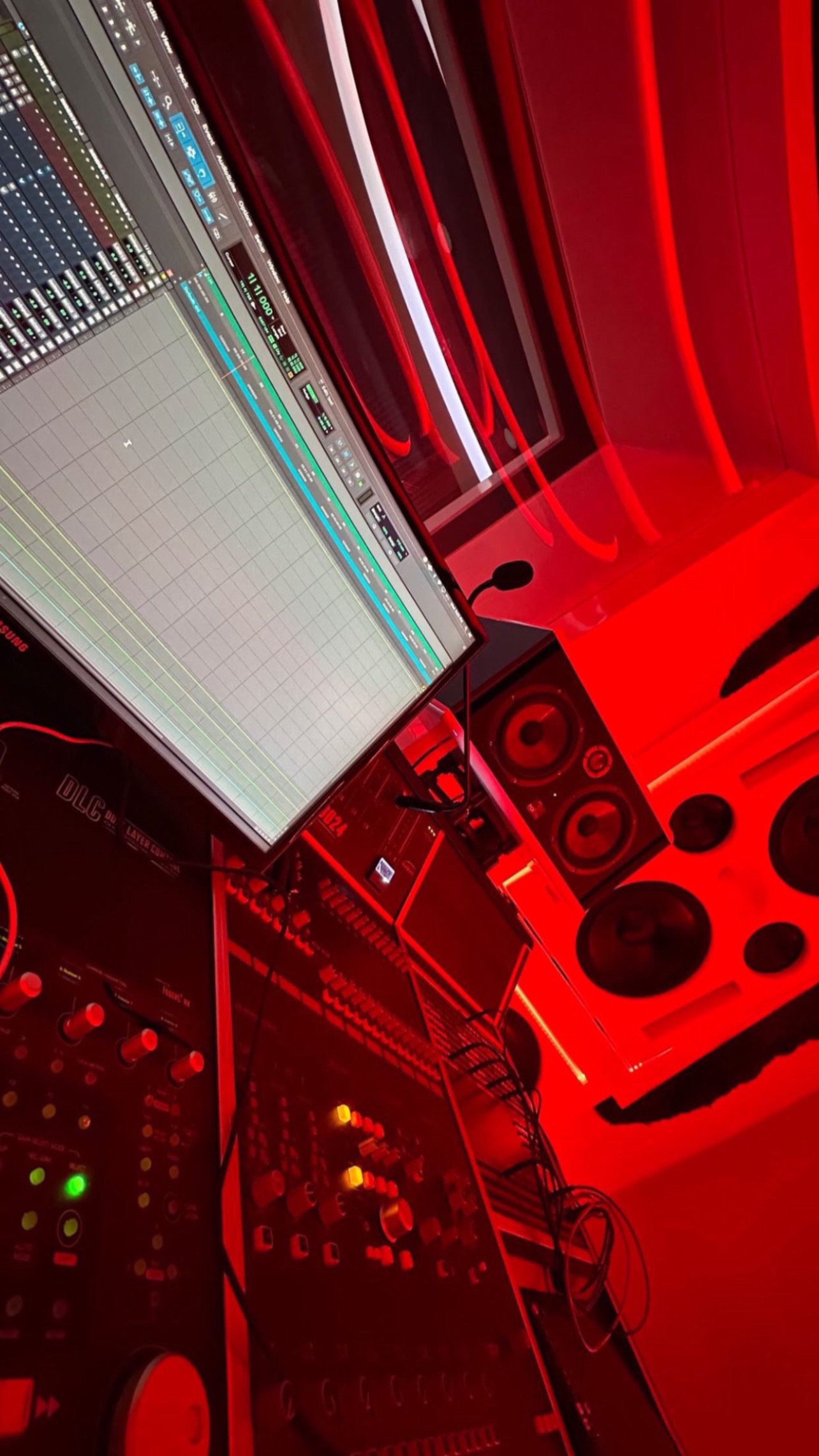 Recording studio with mixing console, computer monitor displaying audio software, and multiple speakers, illuminated by red lighting.