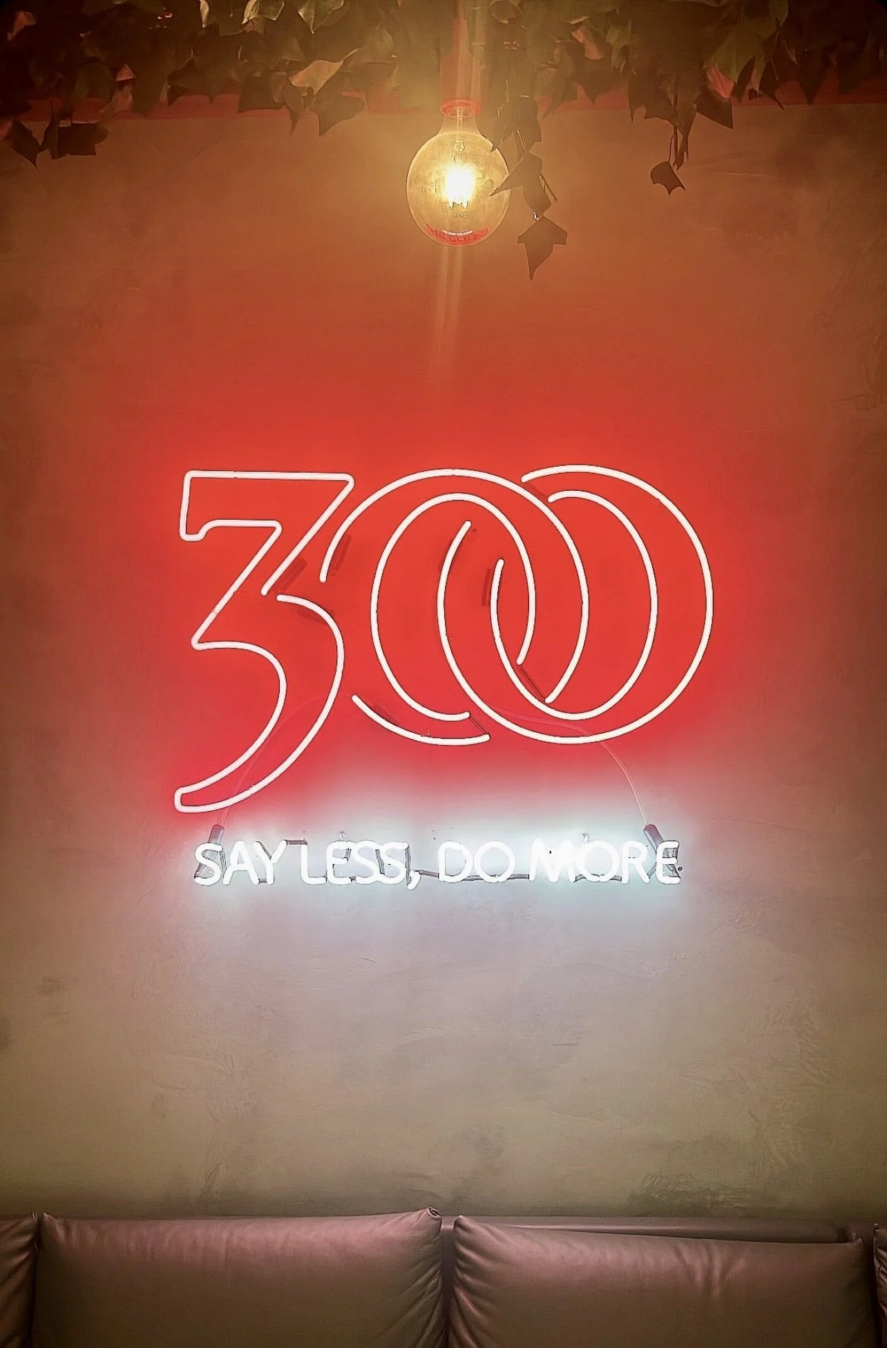 Neon sign reading "300" with overlapping circles and "Say Less, Do More" below, against a gray wall and leather couch, with a hanging bulb above.