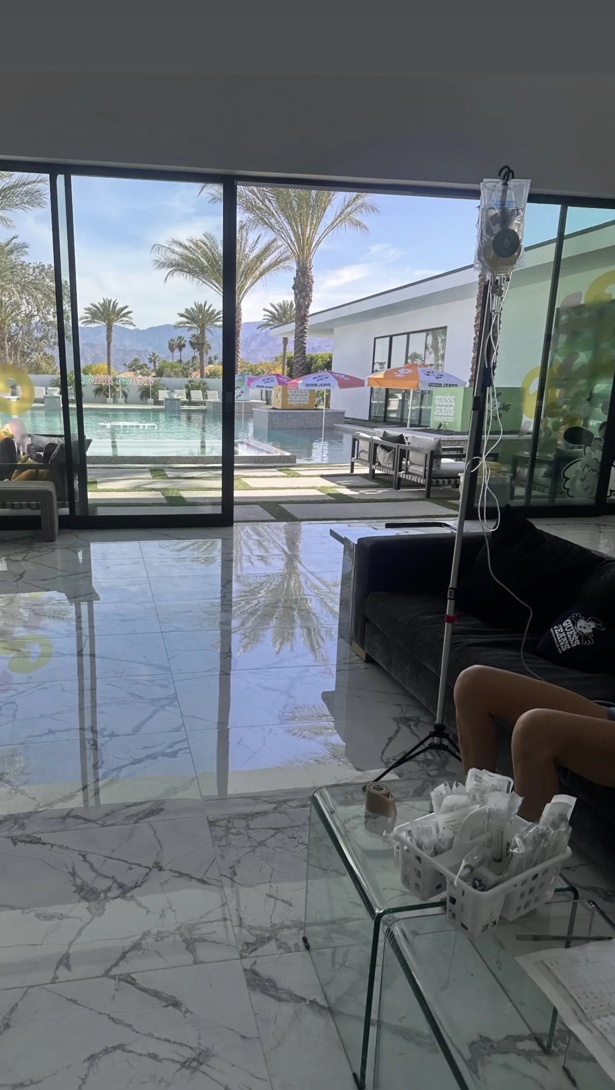 Luxurious living room with glass doors opening to a pool area with palm trees, lounge chairs, and umbrellas. Indoors, there's a marble floor, a black couch with a person seated, a glass table with medical supplies, and a mounted IV drip.