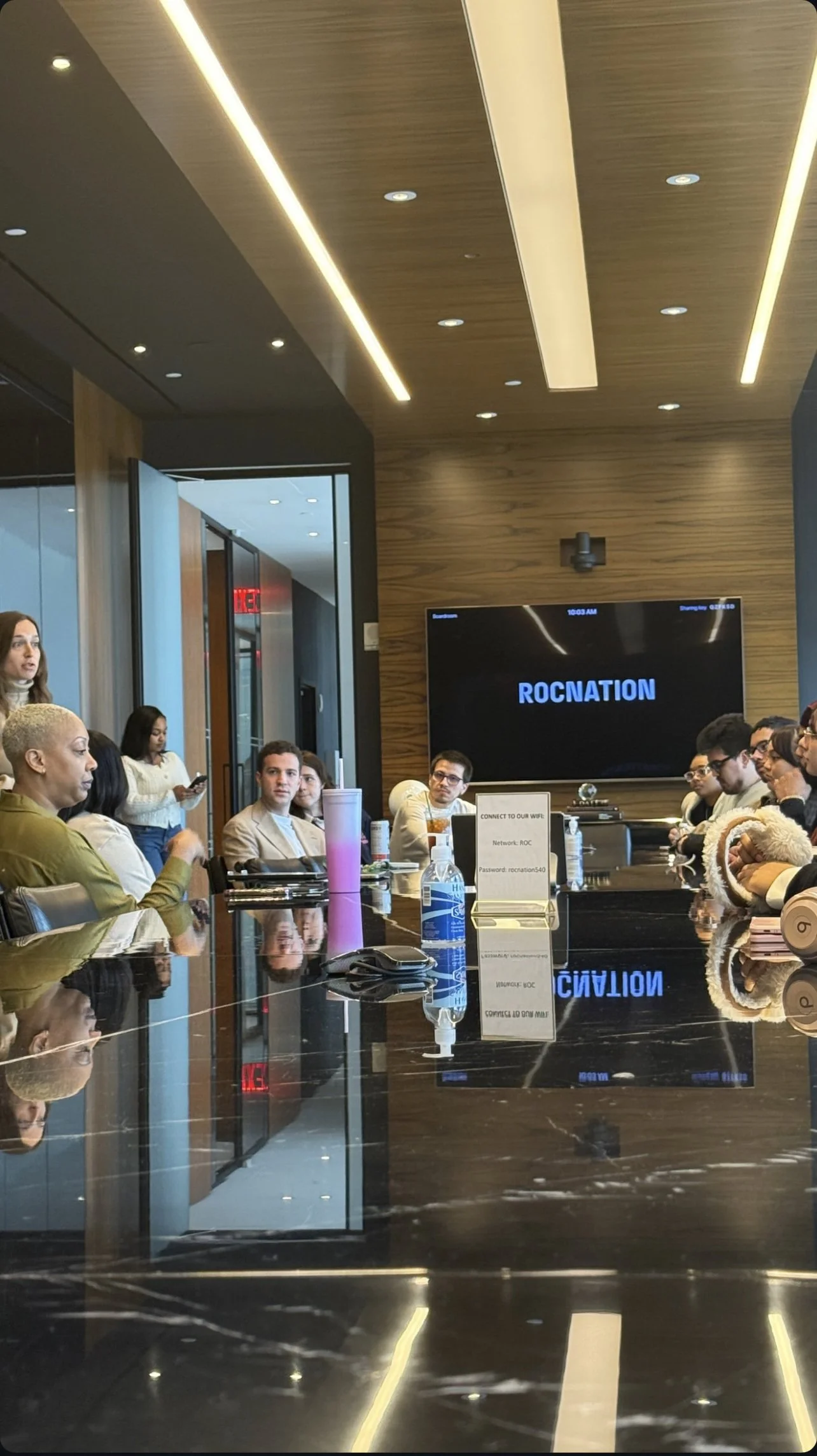 Conference room with people sitting around a large table, a screen displaying 'ROCNATION,' reflective surface, and modern lighting.