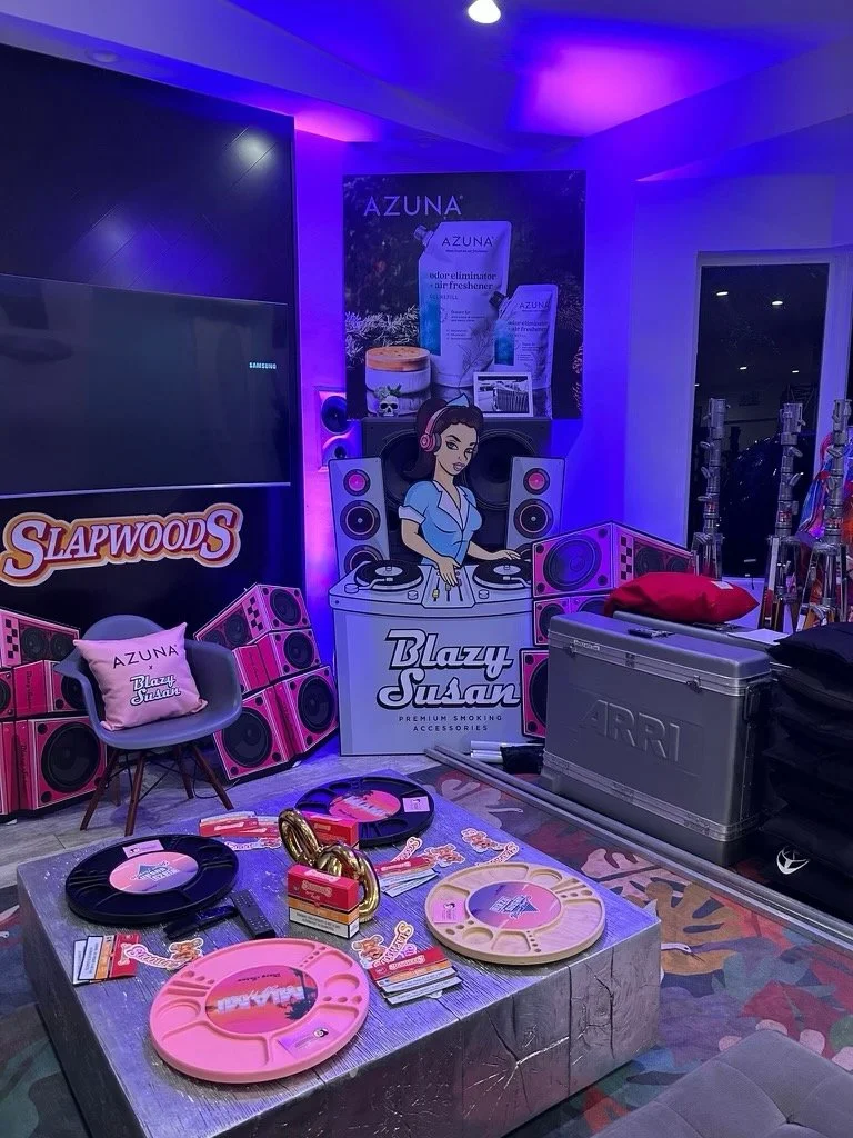 Room with a promotional display featuring Blazy Susan and Azuna products, with pink-themed sound equipment, a chair, hookahs, and a silver ARRI case.