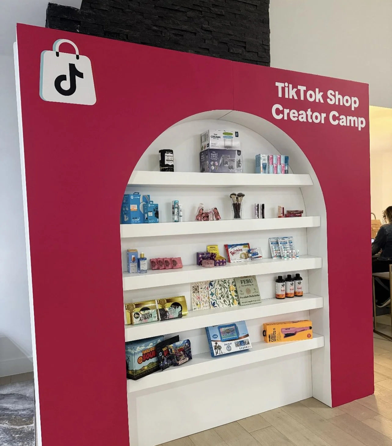 Pink display with "TikTok Shop Creator Camp" text, showcasing various products on shelves including beauty items, healthcare products, and hobby kits. TikTok shopping bag logo on top left.