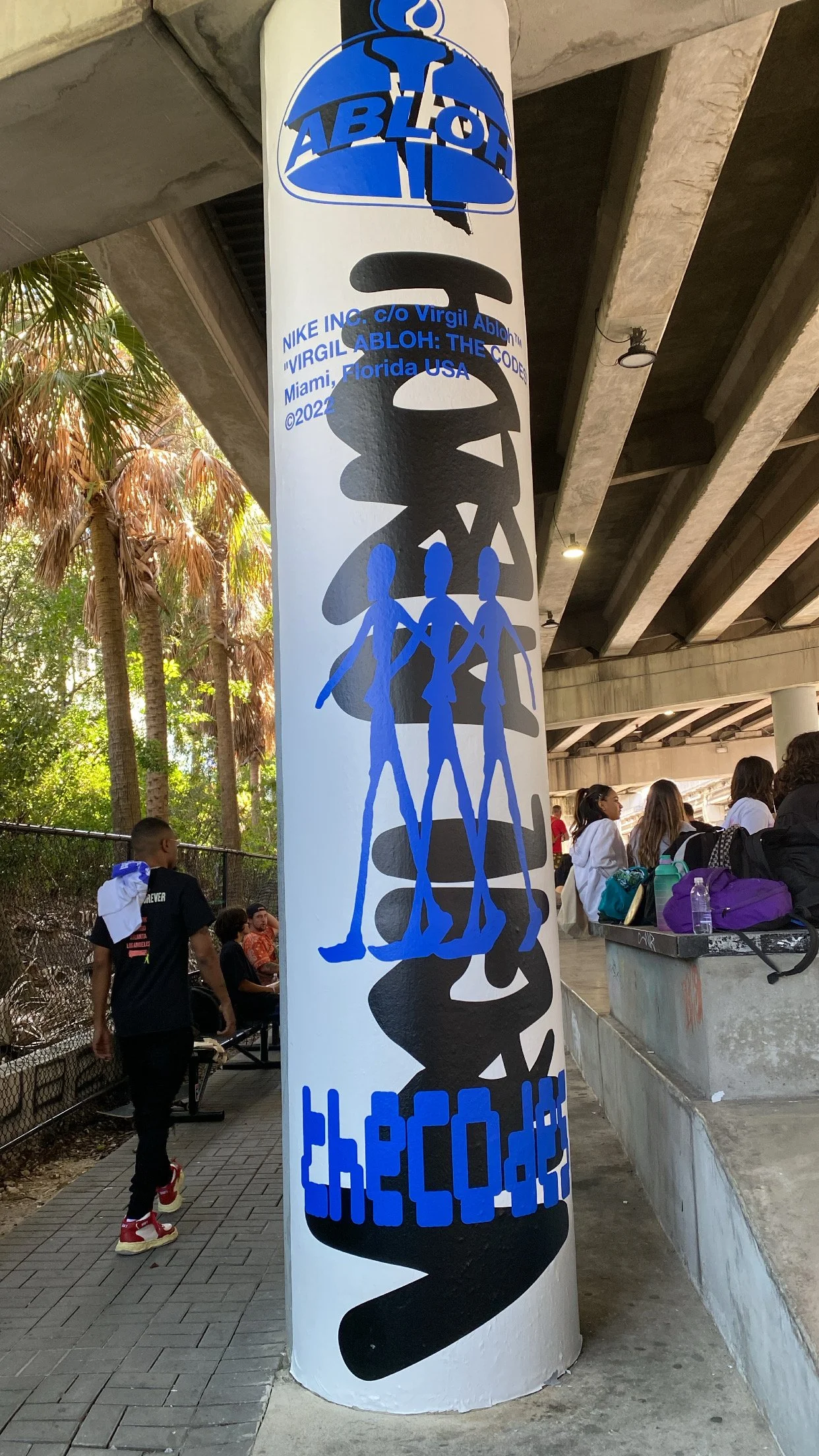 Artistic pillar under a bridge featuring 'ABLOH' and 'theCode' text with abstract blue figures, part of Virgil Abloh exhibition in Miami.