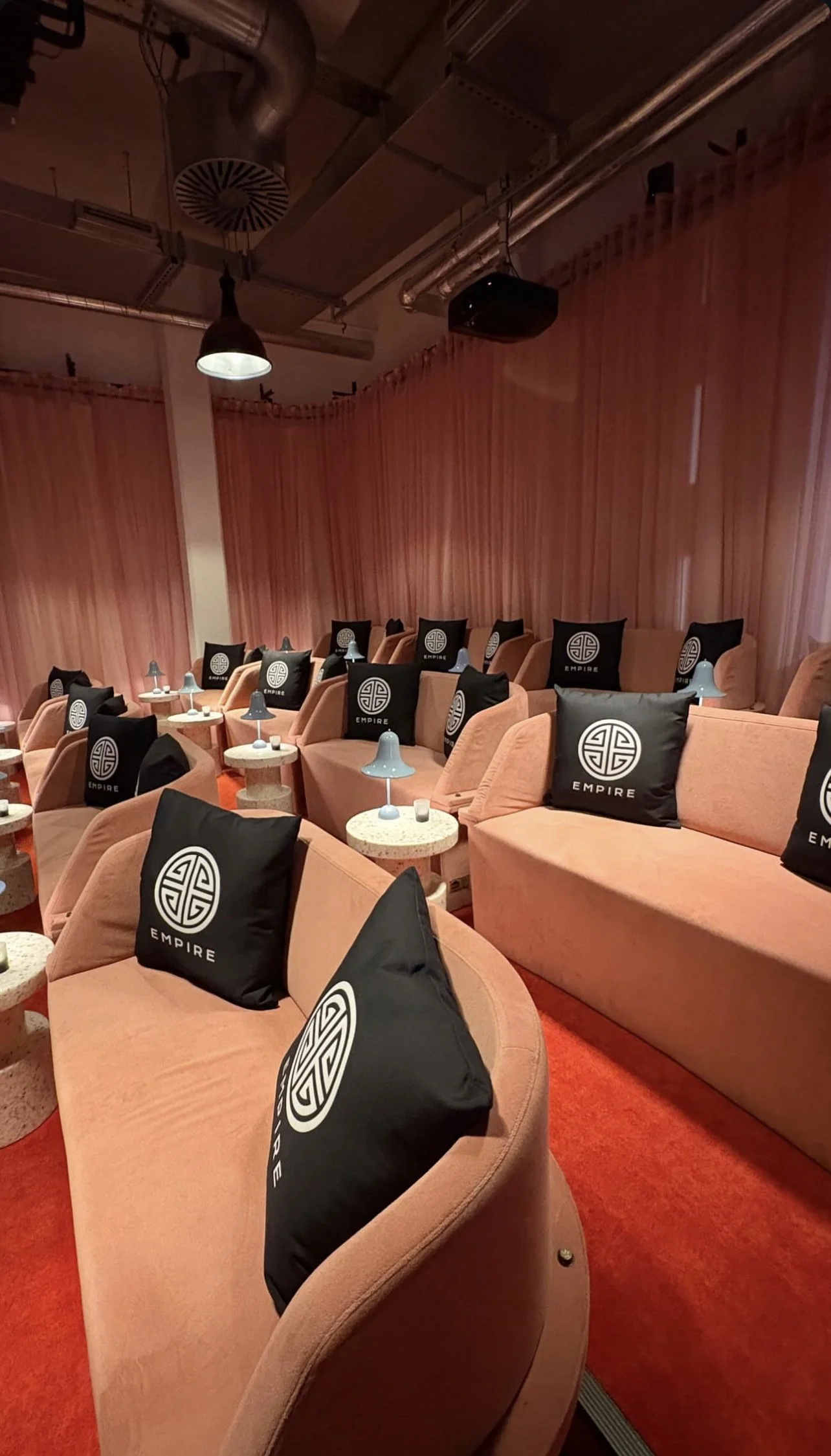 Cozy cinema room with pink seating and black branded cushions, small tables with pastel blue lamps, and dim lighting.