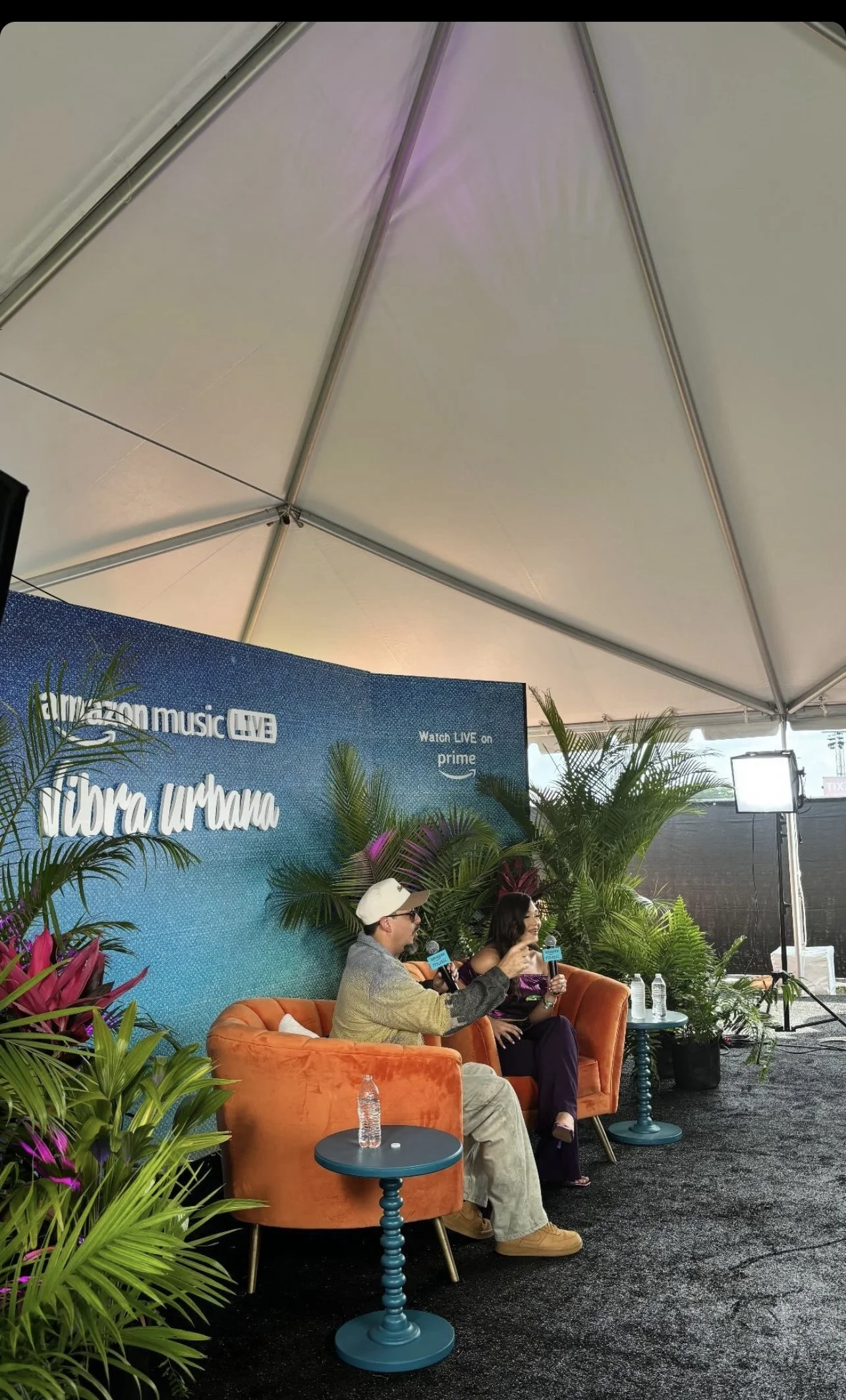 Outdoor interview setup under a white tent featuring two people sitting on orange chairs on a black carpet. A blue backdrop with 'Amazon Music Live' and 'Vibra Urbana' logos is behind them, surrounded by tropical plants. A small round table with a wa