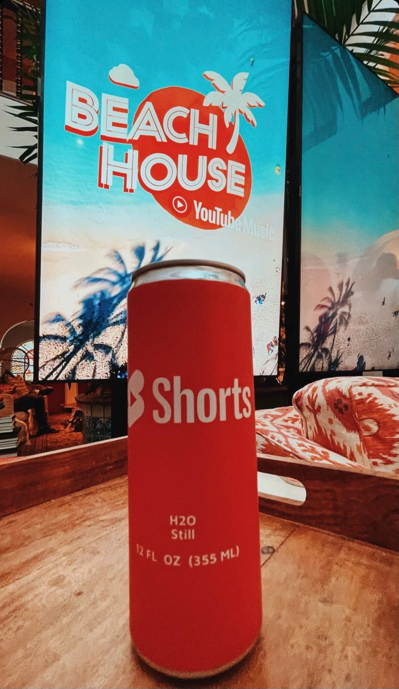 Red can labeled "Shorts H2O Still" on wooden table, with beach-themed screen and palm shadows in the background.
