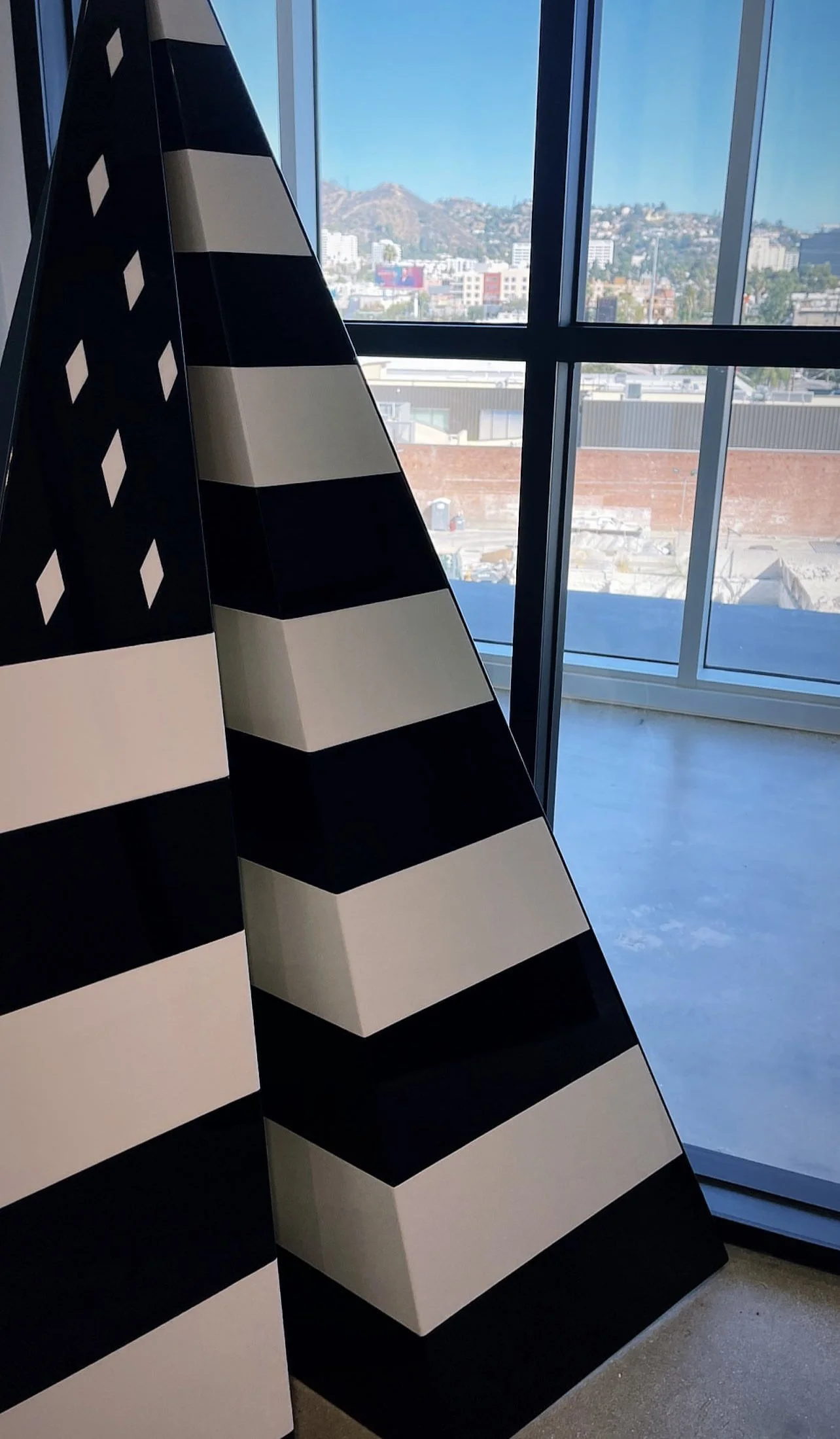 Black and white striped pyramid sculptures inside a room with large windows overlooking a cityscape with hills in the background.