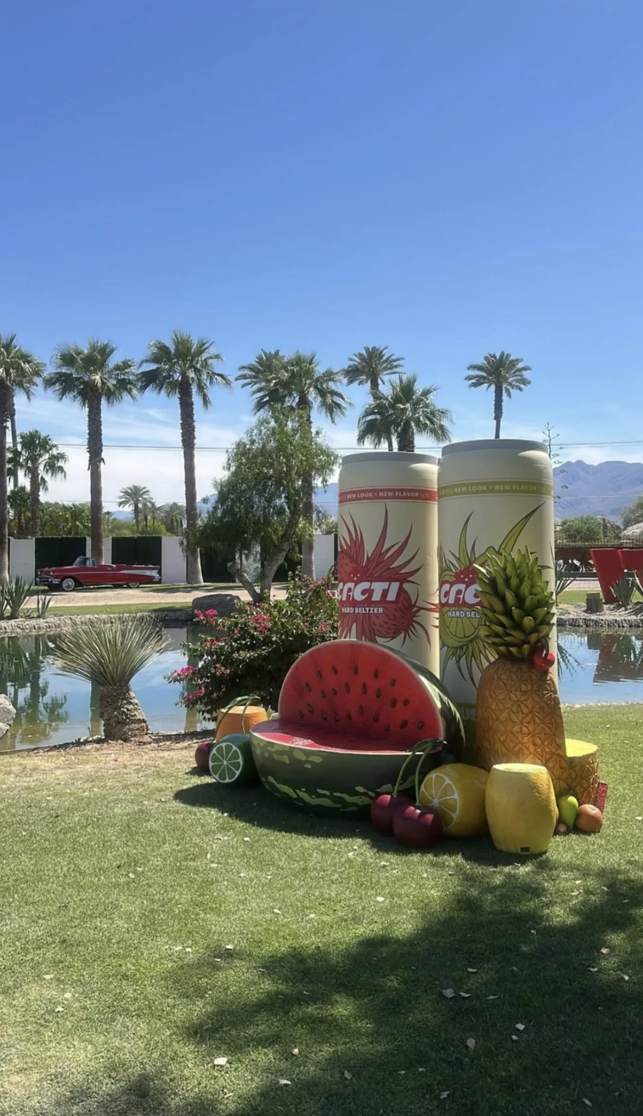 Outdoor scene with large inflatable cans and oversized fruit sculptures, including watermelon, pineapple, cherries, and citrus, near palm trees and a pond.