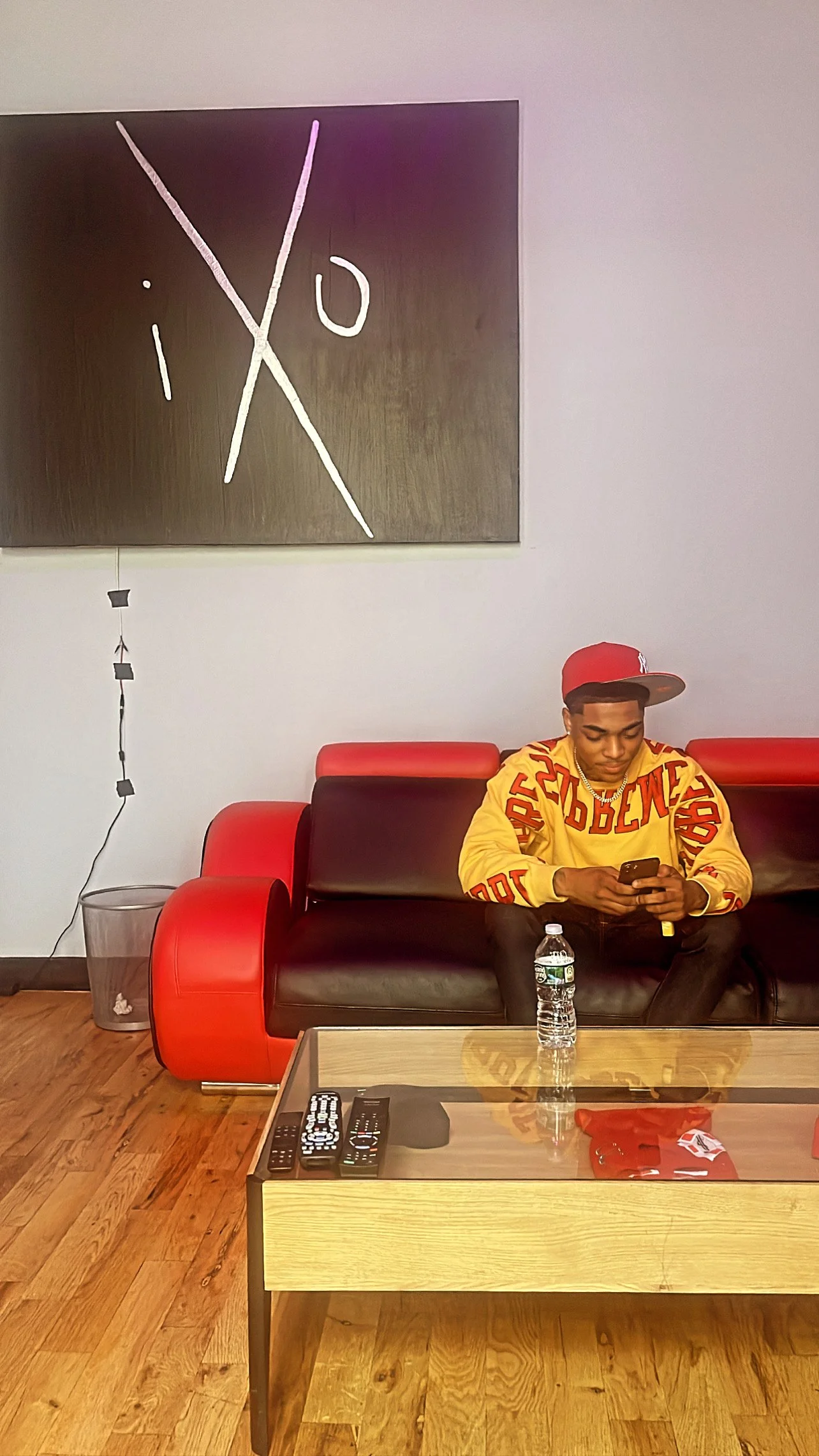 Person sitting on a red and black couch using a smartphone, wearing a yellow sweater and red cap, with a water bottle and TV remotes on a wooden table, under a wall art marked "iXO."