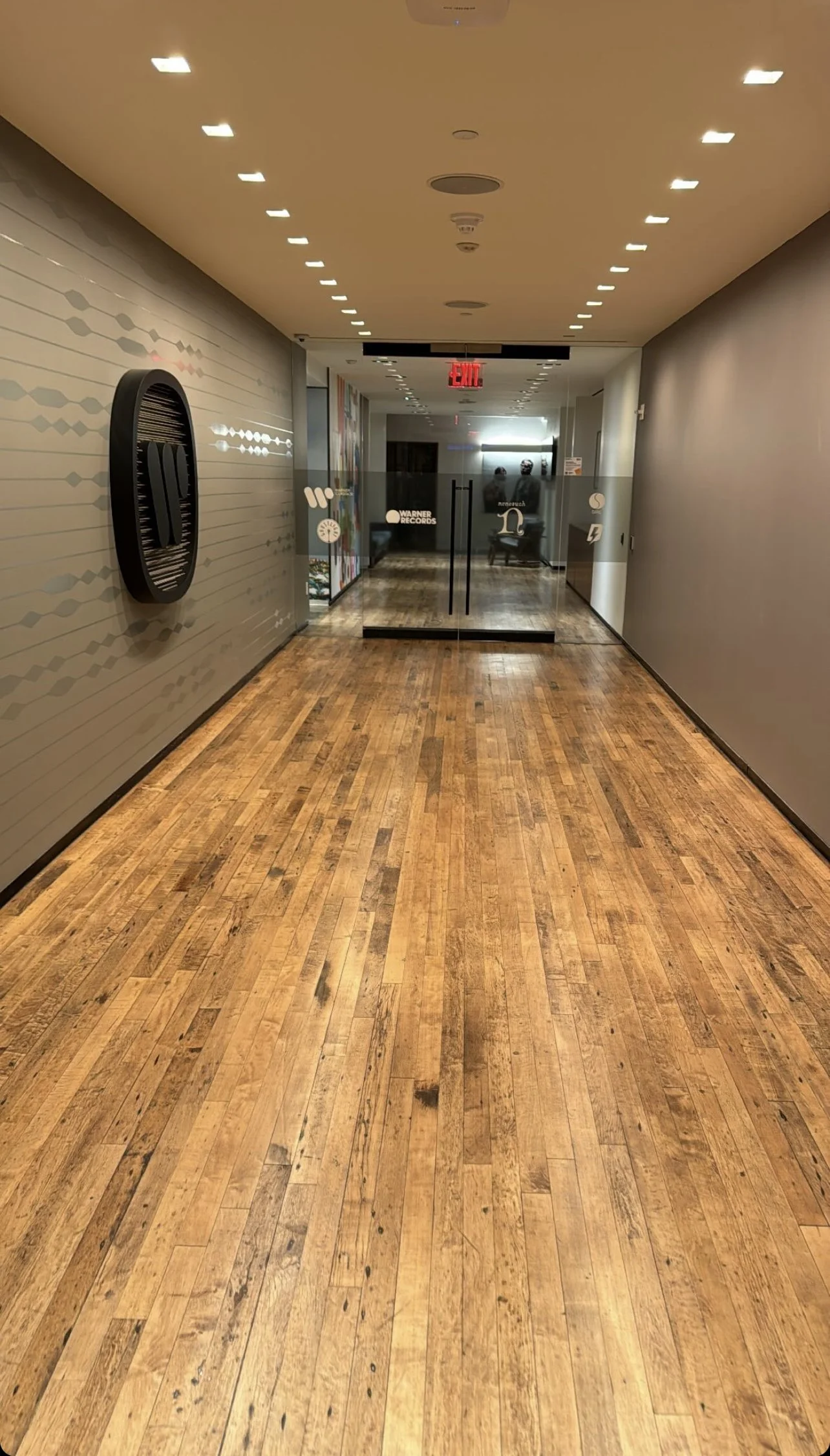 Modern hallway with wooden floor, recessed ceiling lights, mirrored wall, and glass doors featuring logo designs.