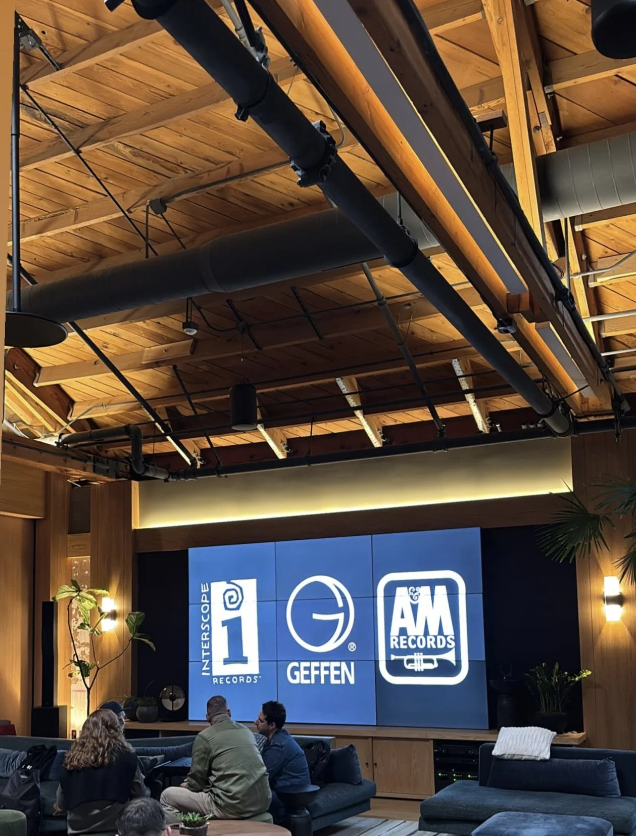 Interior of a modern office lounge with large display showing Interscope Records, Geffen, and A&M Records logos. Ceiling features exposed wooden beams and industrial pipes. People are sitting and conversing on couches.
