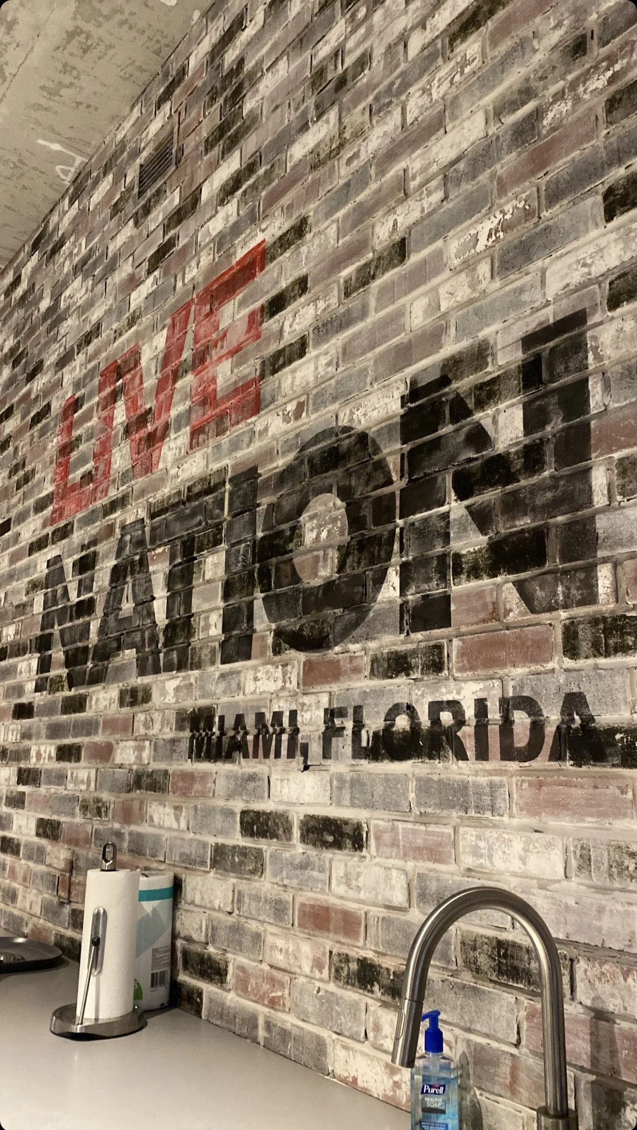 Brick wall with "Live Nation Miami, Florida" text, kitchen counter with paper towel holder, faucet, and hand sanitizer.