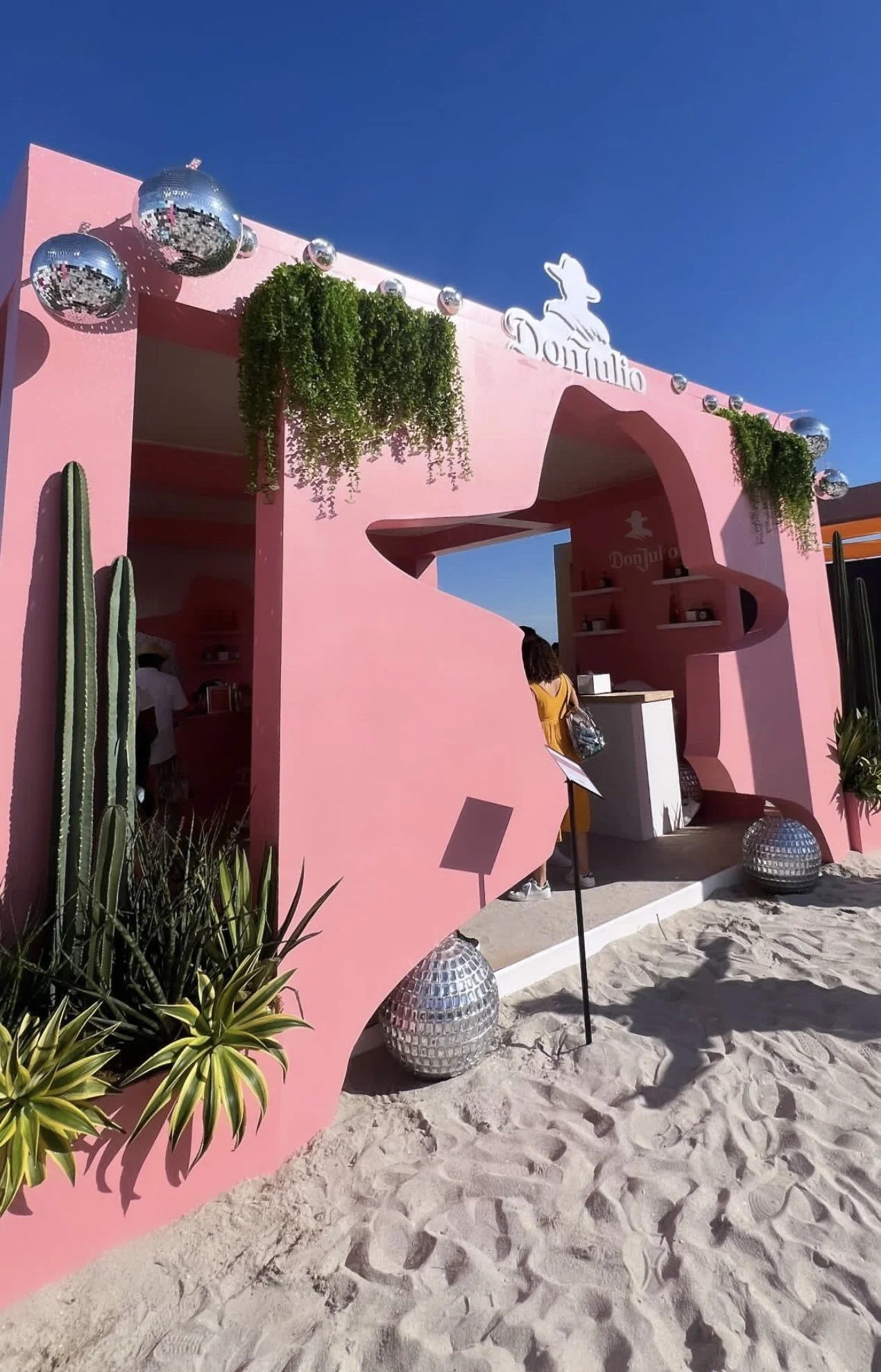 Pink booth set on sand with greenery, silver disco balls, and a cactus, under blue sky.