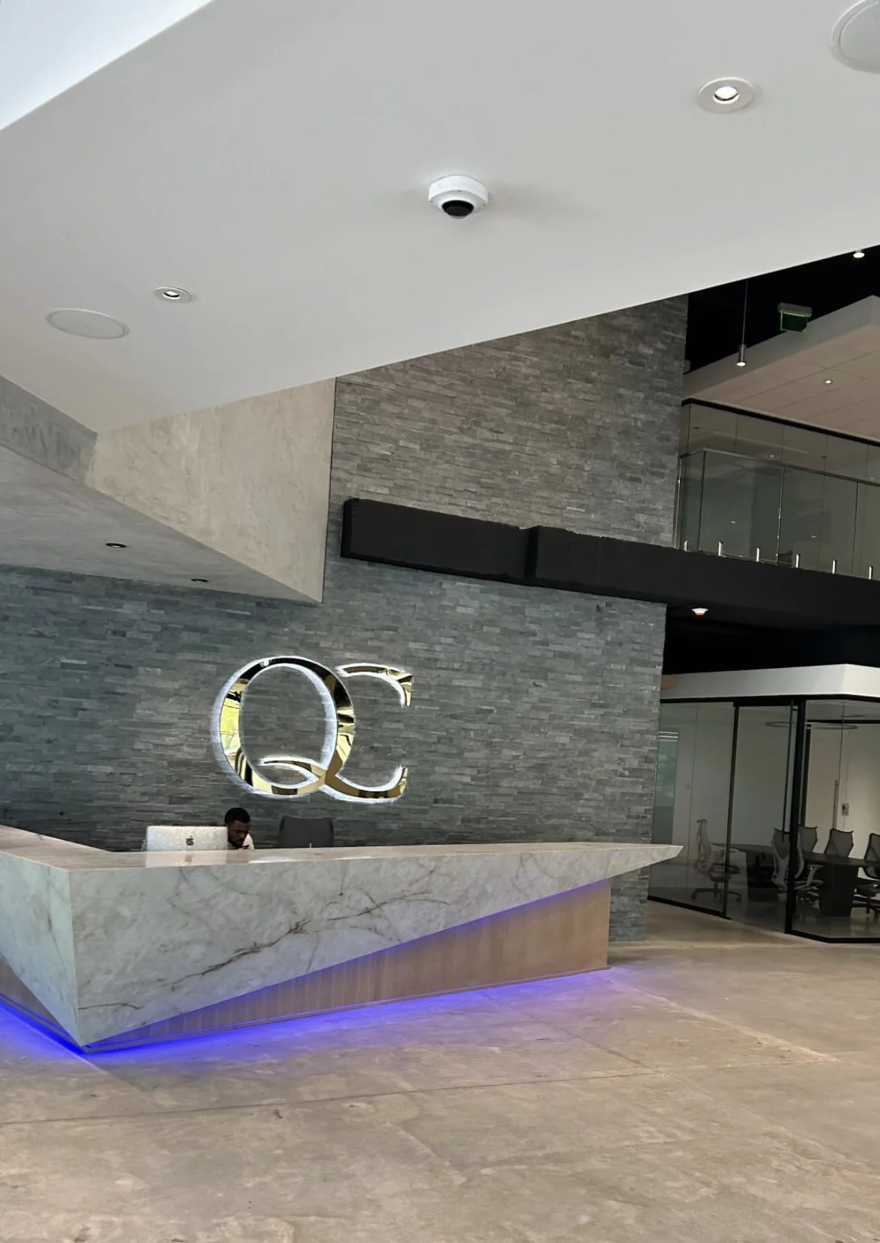 Modern office lobby with gray stone walls, a large reception desk, illuminated blue lighting, and a logo "QC" on the wall.