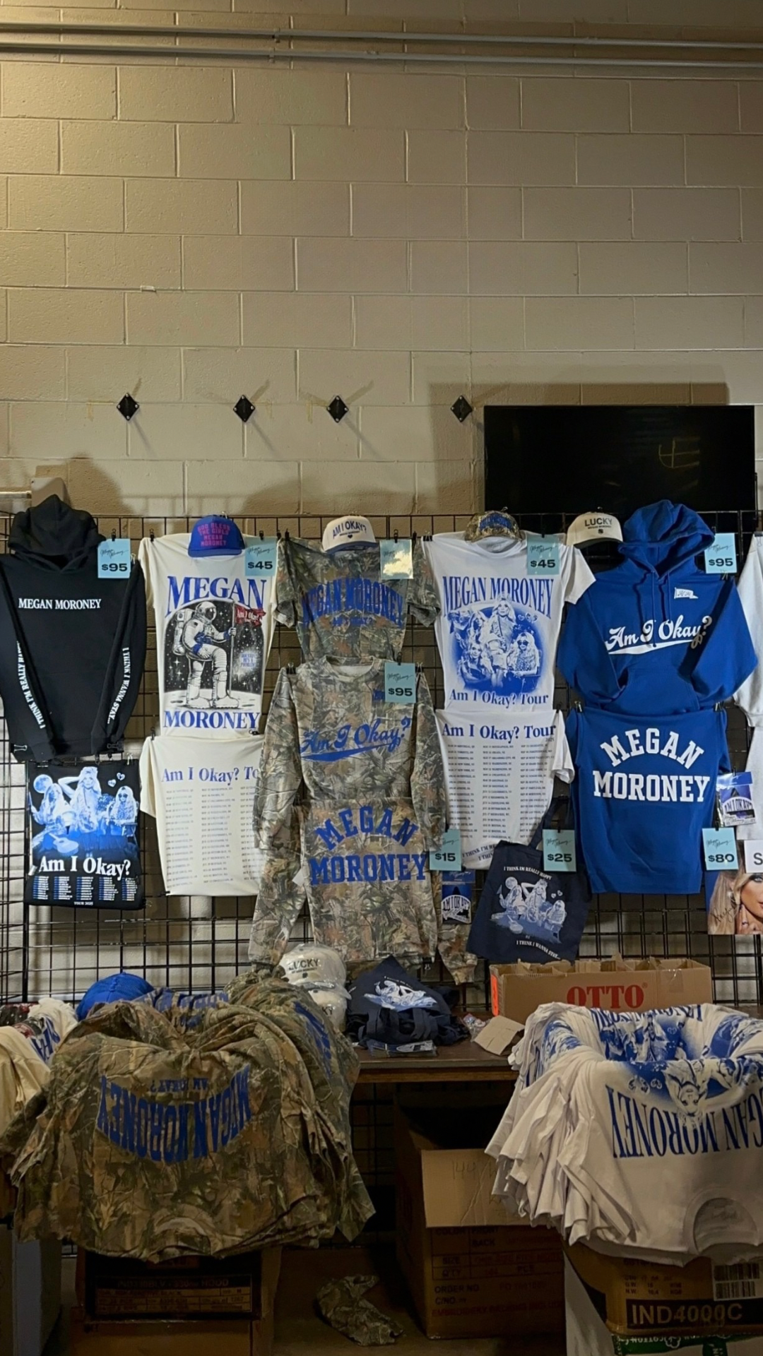Merchandise display for "Am I Okay?" tour with shirts, hoodies, hats featuring Megan Moroney's name and tour graphics.