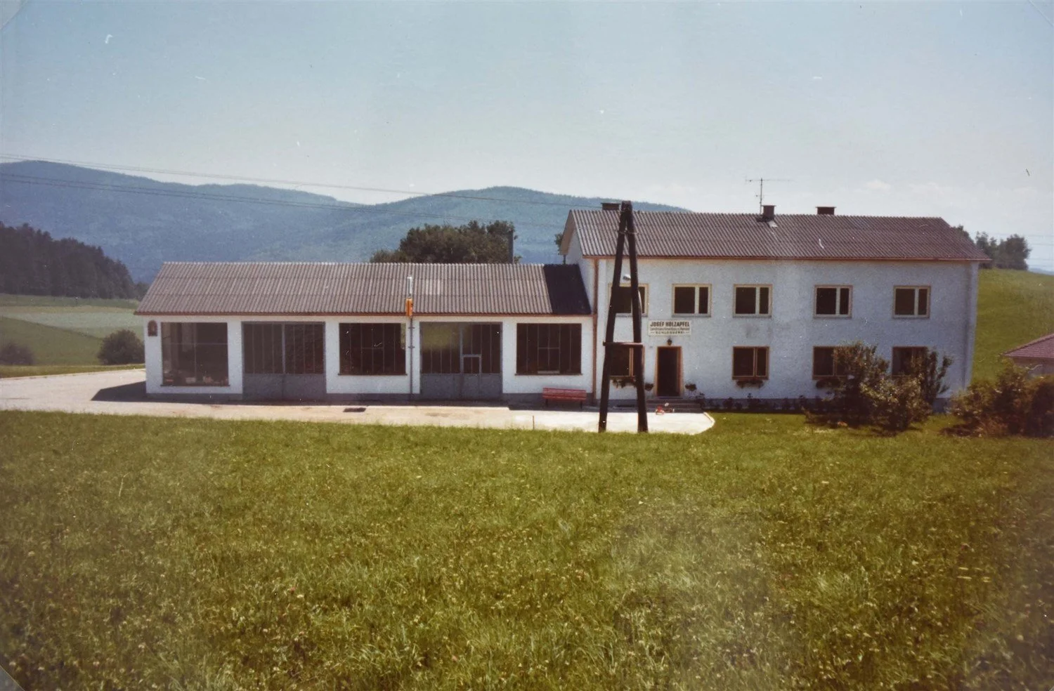Ein zweistöckiges weiße Haus mit roten Ziegeln auf dem Dach, umgeben von grünen Hügeln und Bäumen, bei sonnigem Wetter.