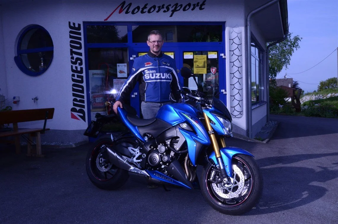 Ein Mann in einem blauen Suzuki-Motorradfahreranzug steht neben einer blauen Suzuki-Motorrad vor einem Gebäude mit dem Wort "Motorsport" an der Fassade.