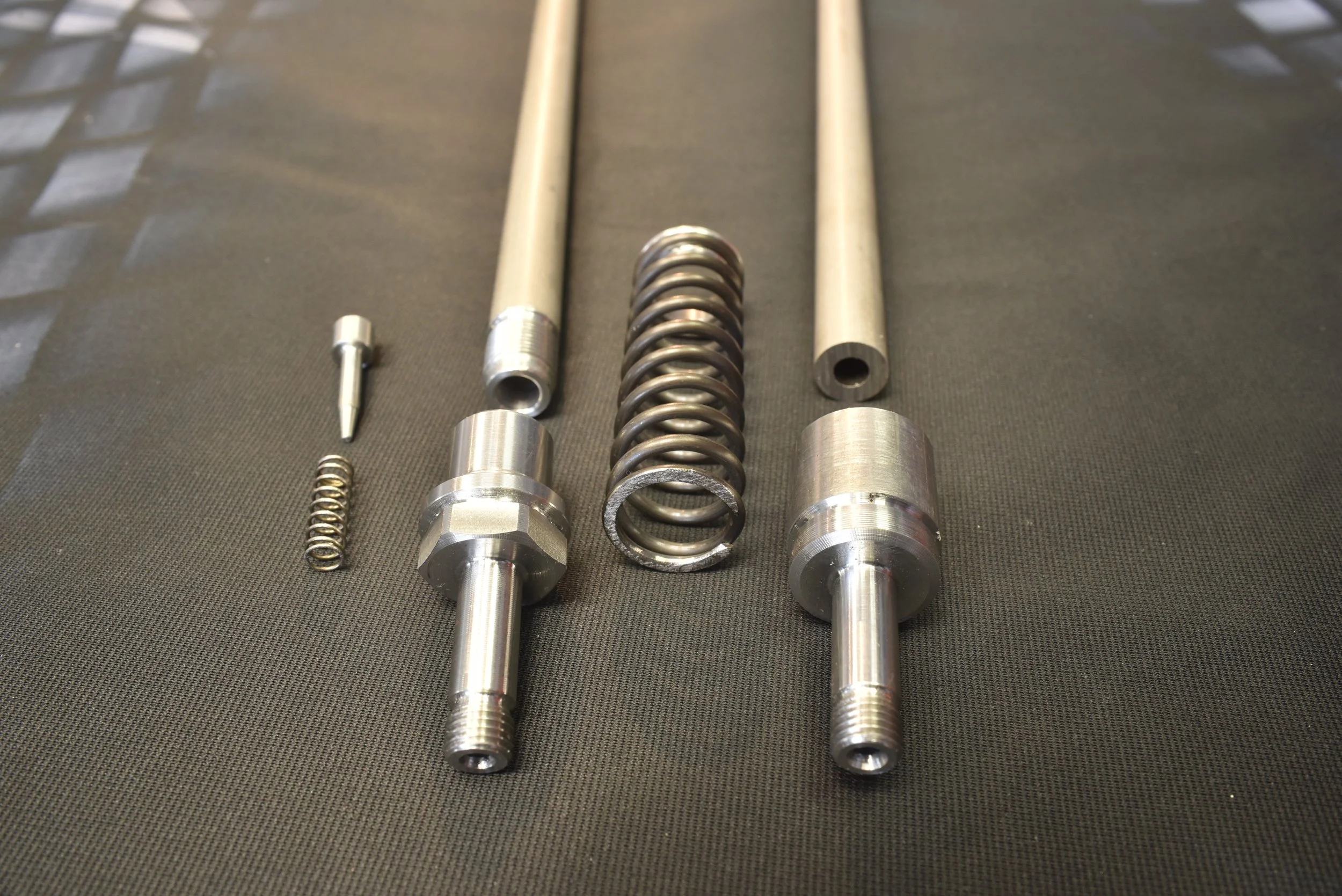 Mehrere metallische Komponenten, darunter Schrauben, Federn und dünne Röhren, auf schwarzem Hintergrund.