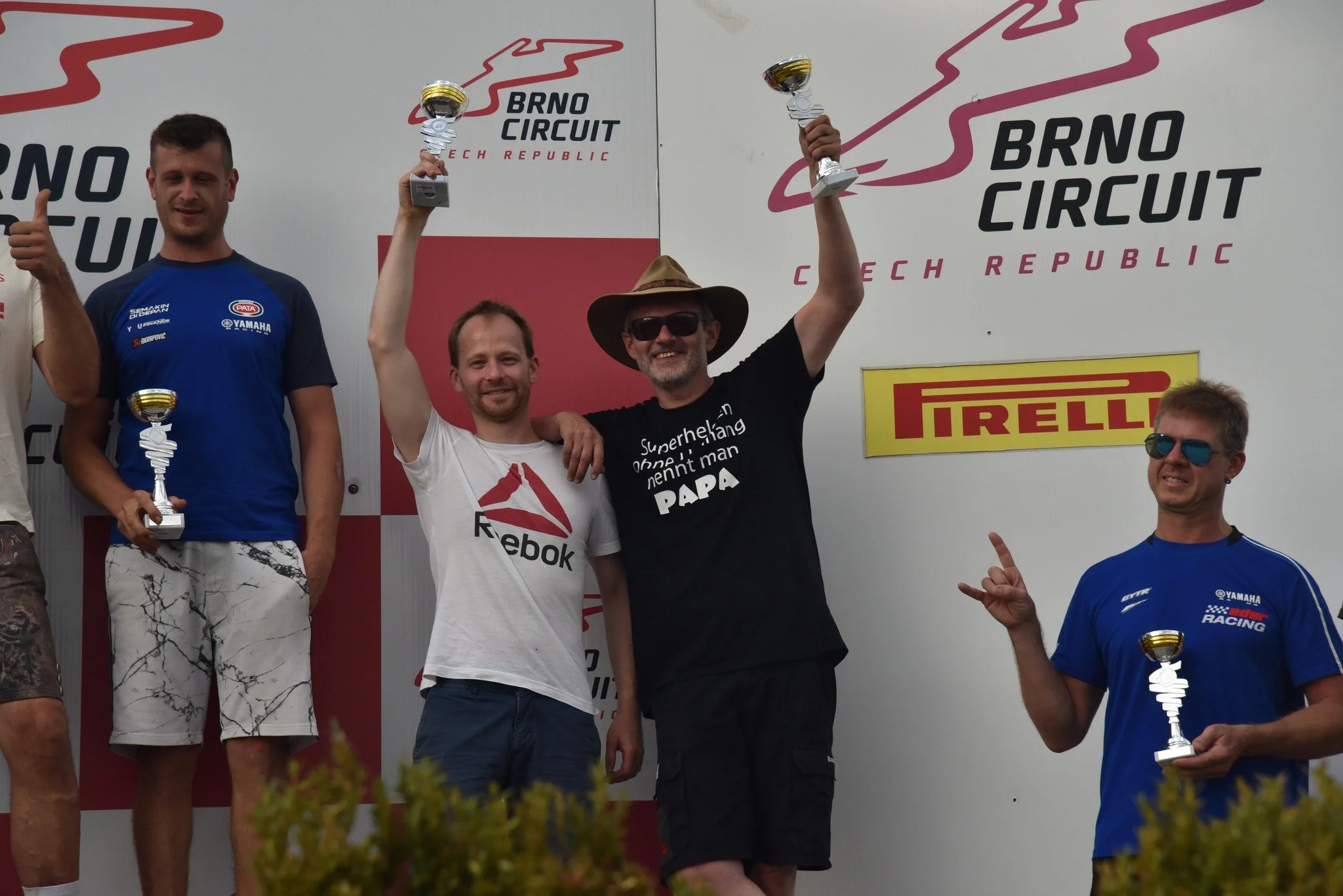 Vier Männer stehen auf der Siegerpodest beim Brno Grand Prix in Tschechien. Zwei Männer halten Pokale hoch, der Mann in der Mitte trägt eine Sonnenbrille und einen Hut, er freut sich und lächelt. Der Mann auf der rechten Seite hält einen Pokal, trägt eine blaue Rennfahrerjacke und Sonnenbrille, und deutet mit der Hand nach oben. Im Hintergrund ist die Brno Circuit Logo und das Pirelli Logo sichtbar.