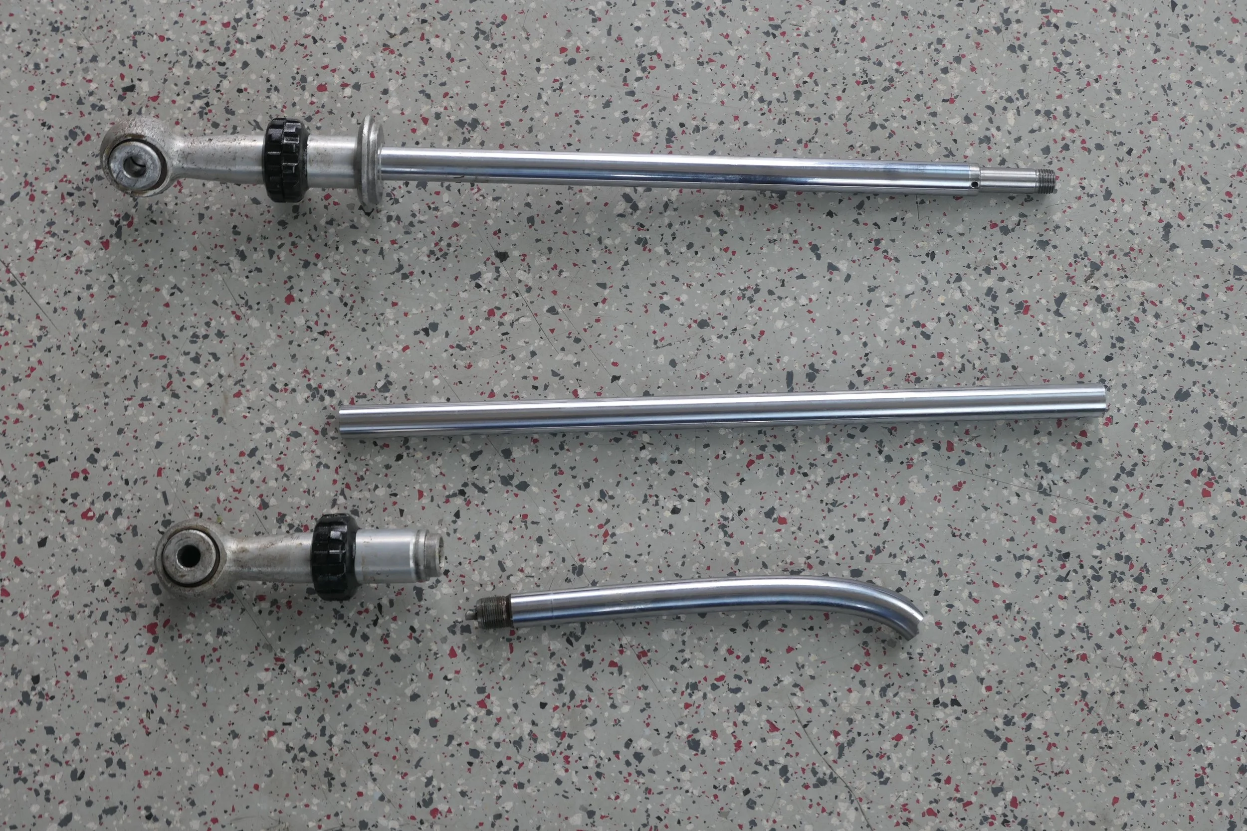 Drei Metallteile, vermutlich für ein Fahrrad oder Motorrad, auf einem grauen, gepunkteten Boden. Das oberste Teil ist eine Gabel mit einem verstellbaren schwarzen Ring, das mittlere ist ein langes, glattes Metallrohr, und das unterste ist ein gebogen