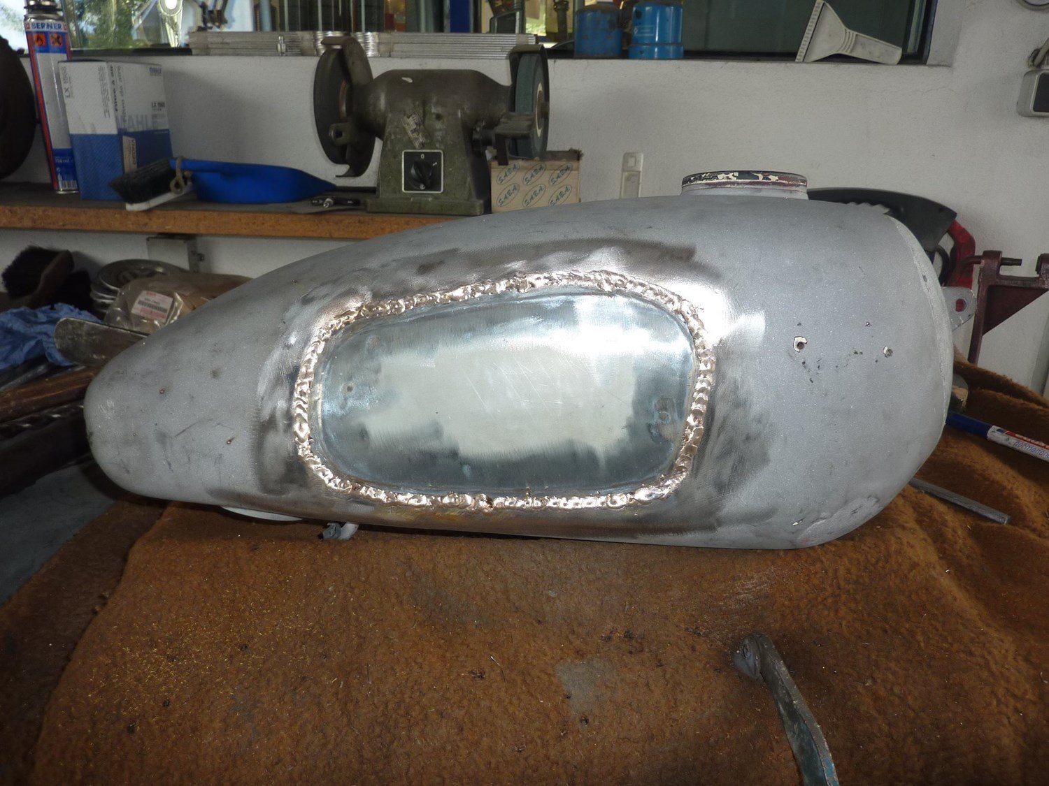 Blick auf ein metallgehendes Fahrzeugtank in einer Werkstatt, umgeben von Werkzeugen, Schrott und anderen Werkstücken.