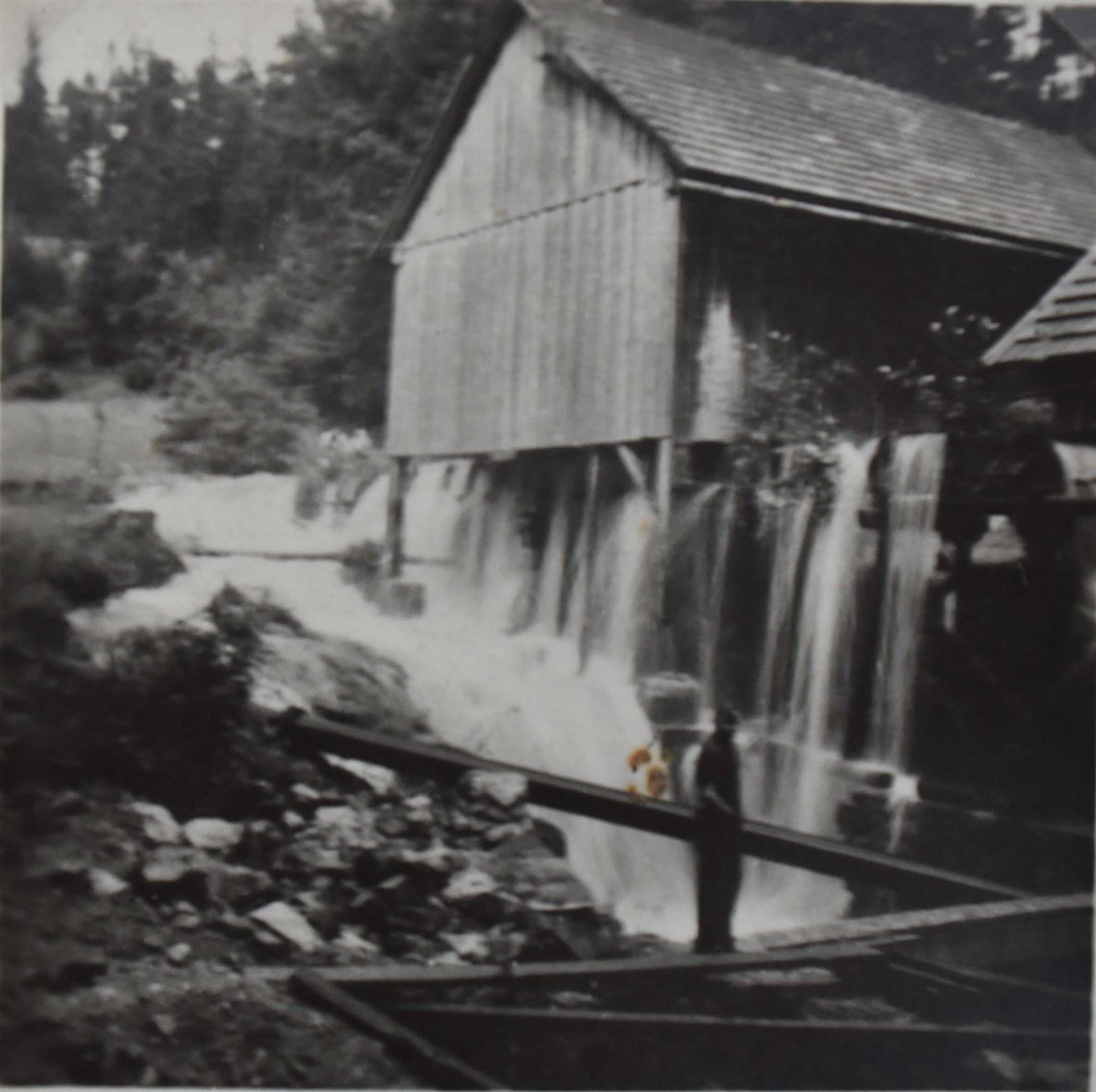 Schwarz-weiß-Foto eines alten Wassermills mit Wasser, das über eine Dammstruktur fließt.