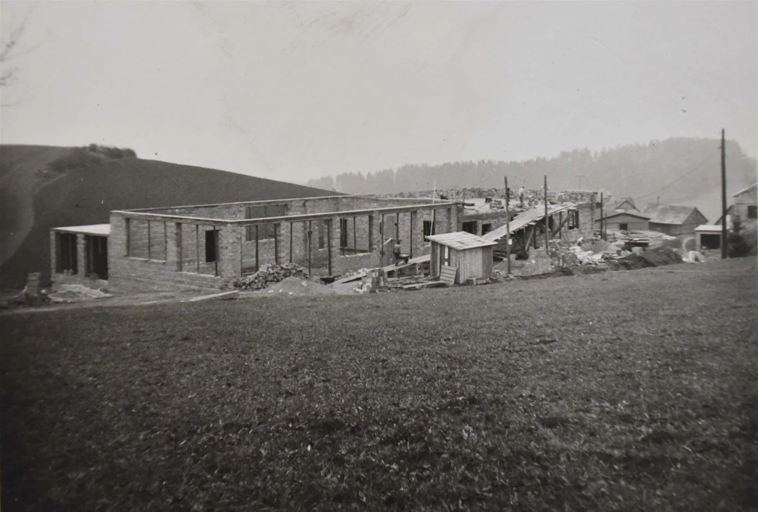 Schwarz-Weiß-Foto eines im Bau befindlichen Gebäudes in ländlicher Umgebung mit Hügeln im Hintergrund, einige Bauarbeiter und Baumaterialien sind sichtbar.