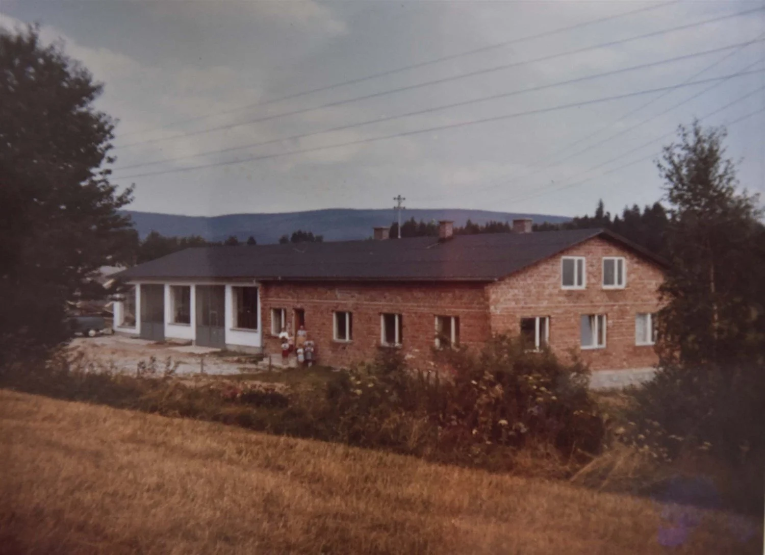 Ein zweistöckiges Haus, teils aus rotem Ziegelstein, mit mehreren Fenstern und einem dunklen Dach, umgeben von Bäumen und einem Rasen, im Hintergrund Berge und ein bewölkter Himmel.