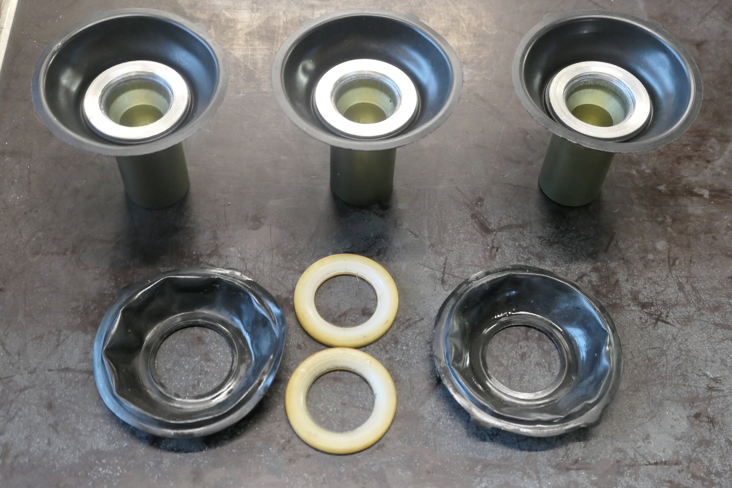 Drei Metallteile mit Gummidichtungen, zwei Gummiringe, und zwei Metallbuchsen auf einer Metalloberfläche.