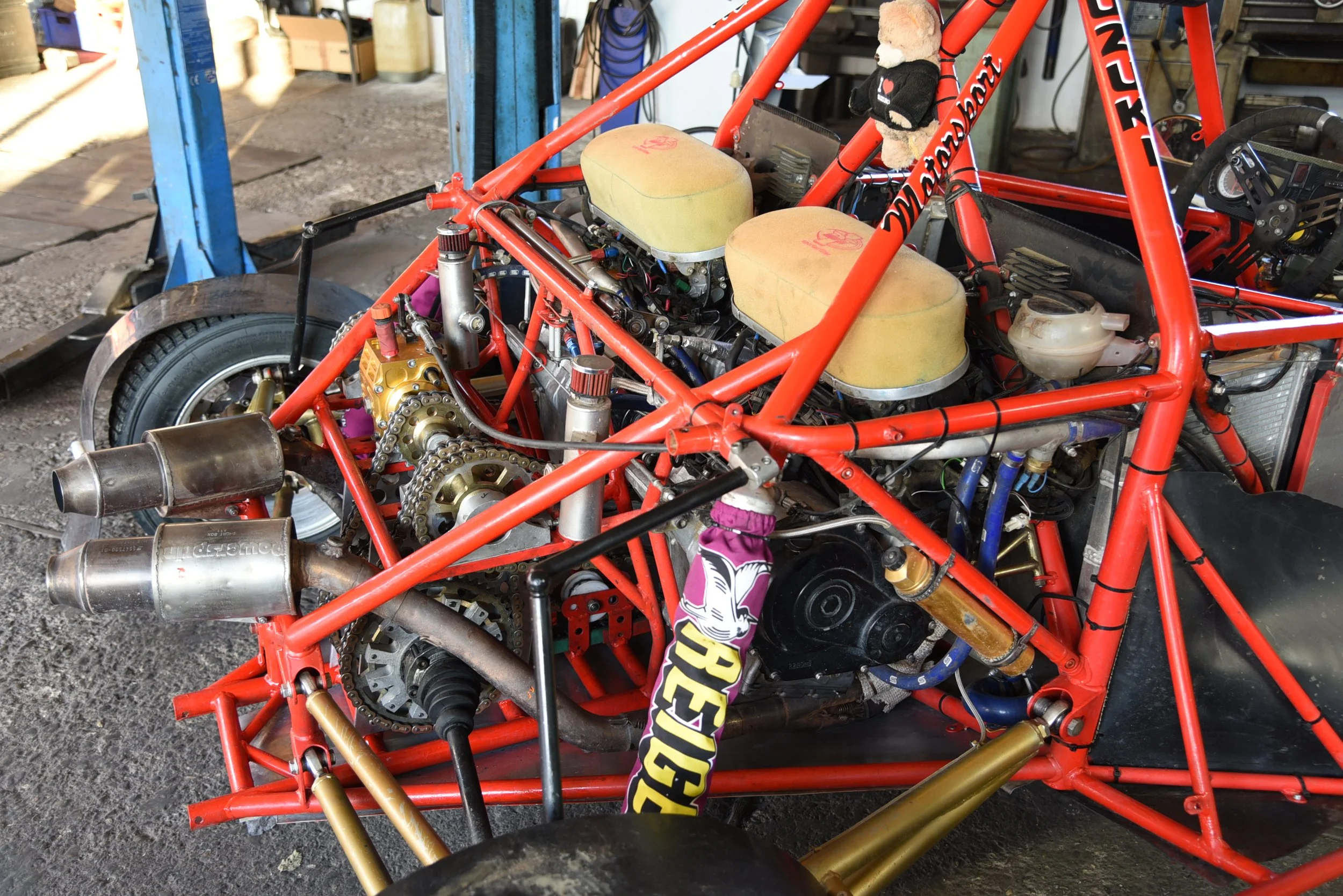 Innenansicht eines Rennwagens mit rotem Rahmen, sichtbarem Motor, Auspuffanlagen und verschiedenen technischen Komponenten in einer Werkstatt.