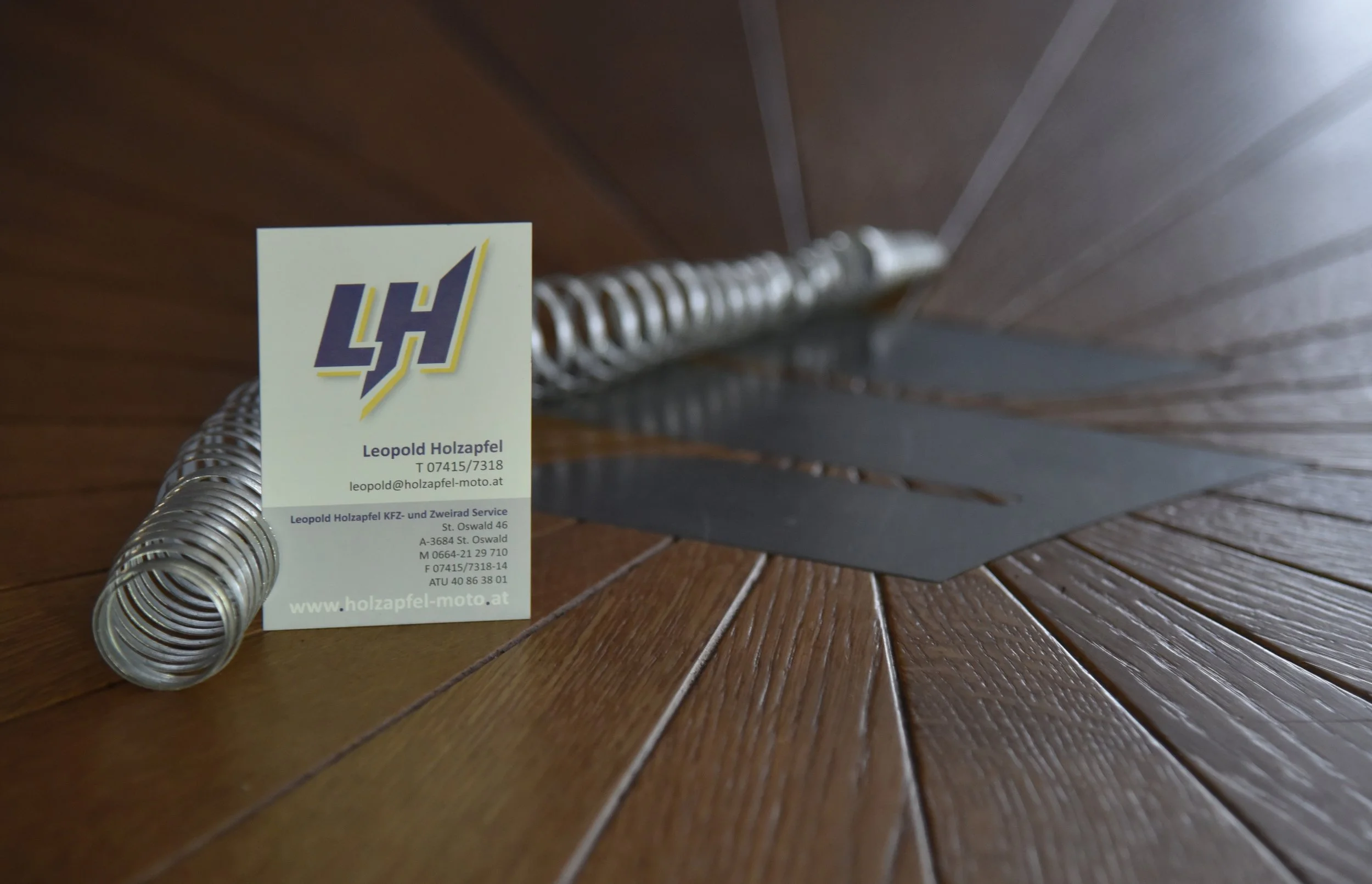 Geschäftskarte auf Holzdielen neben einem Metallspiralrohr. Die Karte gehört Leopold Holzapfel, einem KFZ- und Zweiradservice. Die Webseite lautet www.holzapfel-moto.at.