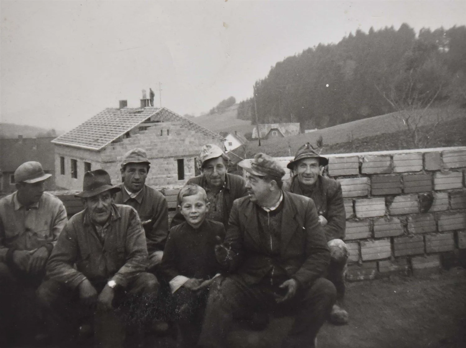 Sieben Männer und ein Junge sitzen vor einem gemauerten Zaun bei einem Häuserbau im ländlichen Gebiet, vermutlich in der Vergangenheit, schwarz-weiß, mit Hüten und Arbeitkleidung.