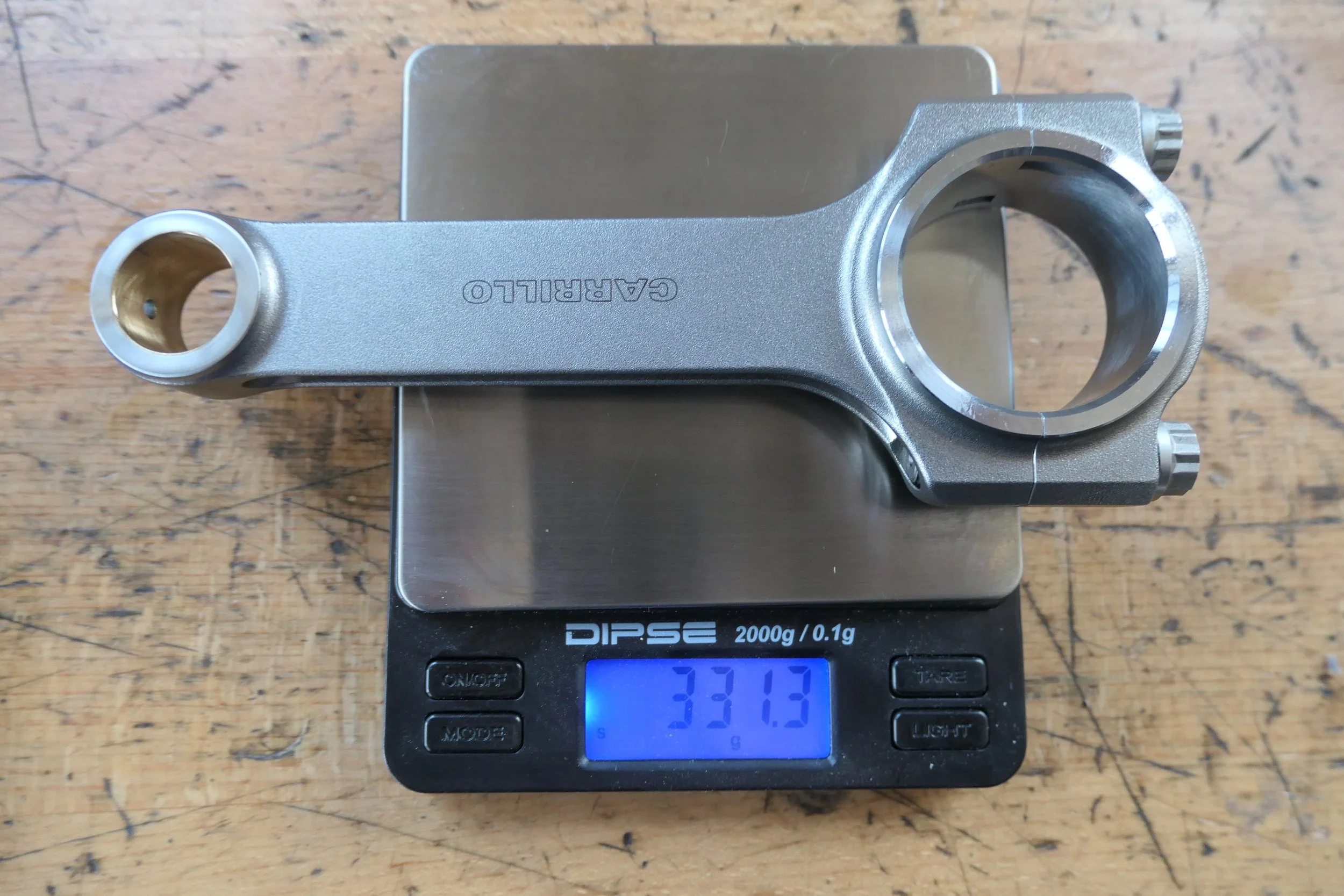 Ein Fahrradsteuerkopfbügel aus Metall, der auf einer digitalen Waage mit 331,3 g Gewicht liegt, auf einem Holztisch.