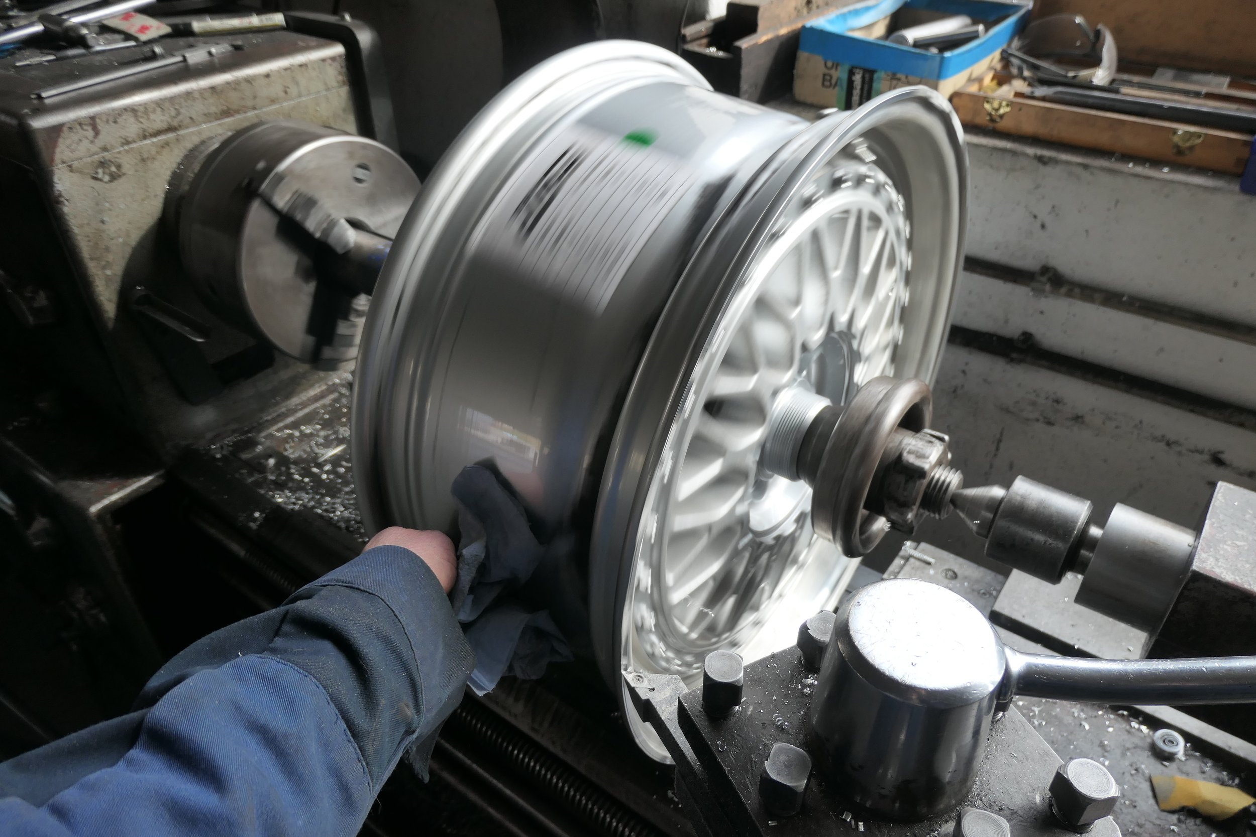 Ein Mechaniker bearbeitet ein großes, silbernes Felgenrad auf einer Werkbank. Im Hintergrund sind Werkzeuge und Maschinen sichtbar.