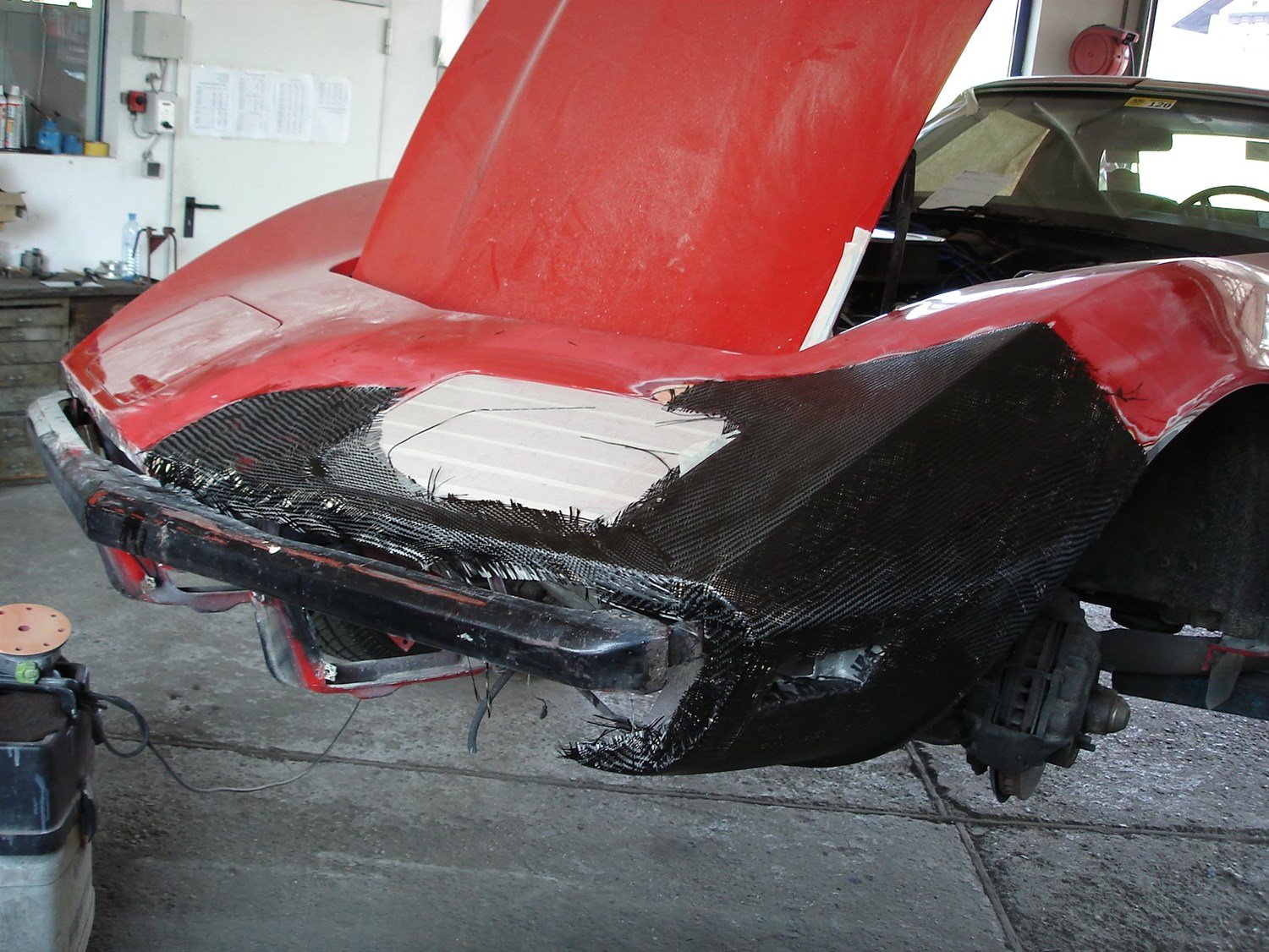 Autowrack in einer Werkstatt, bei dem die vordere Stoßstange entfernt wurde und die Karosserie mit schwarzem Material hinterlegt ist.