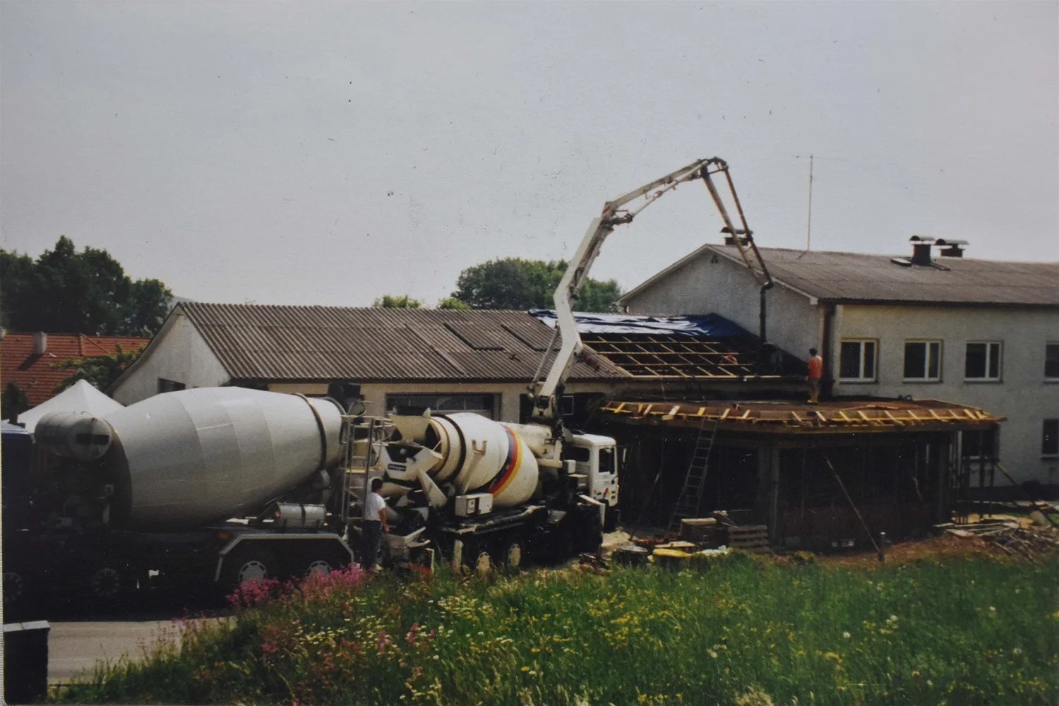 Bauarbeiten an einem Wohnhaus, mit einem Kran, der Dachziegel verlegt, und einem Betonmischer auf dem Gelände.