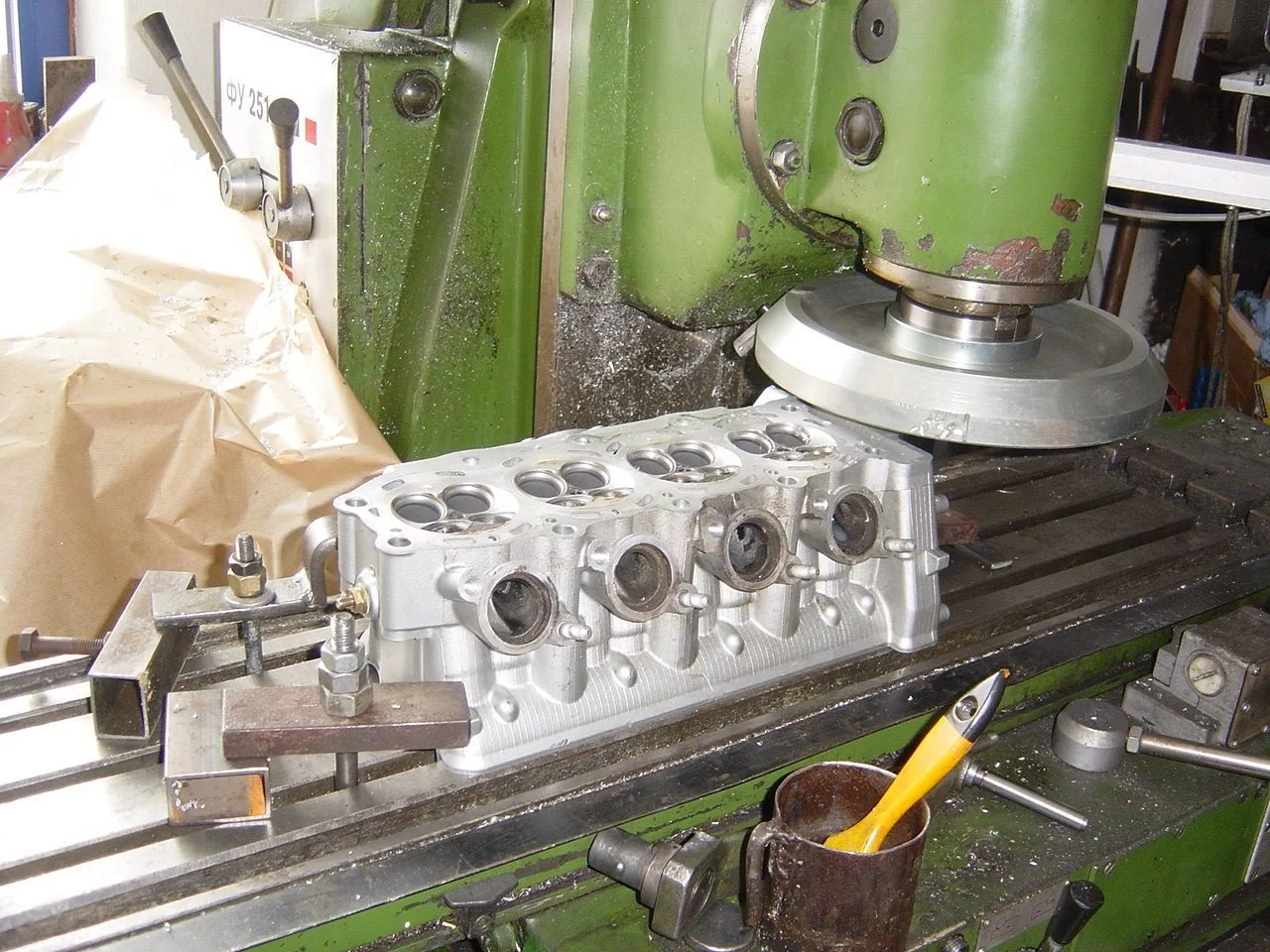 Eine Werkstatt mit einer grünen Maschine, die eine Aluminiummotorhaube bearbeitet, sowie ein Metallrahmen und ein Pinsel in einem Behälter auf der Werkbank.