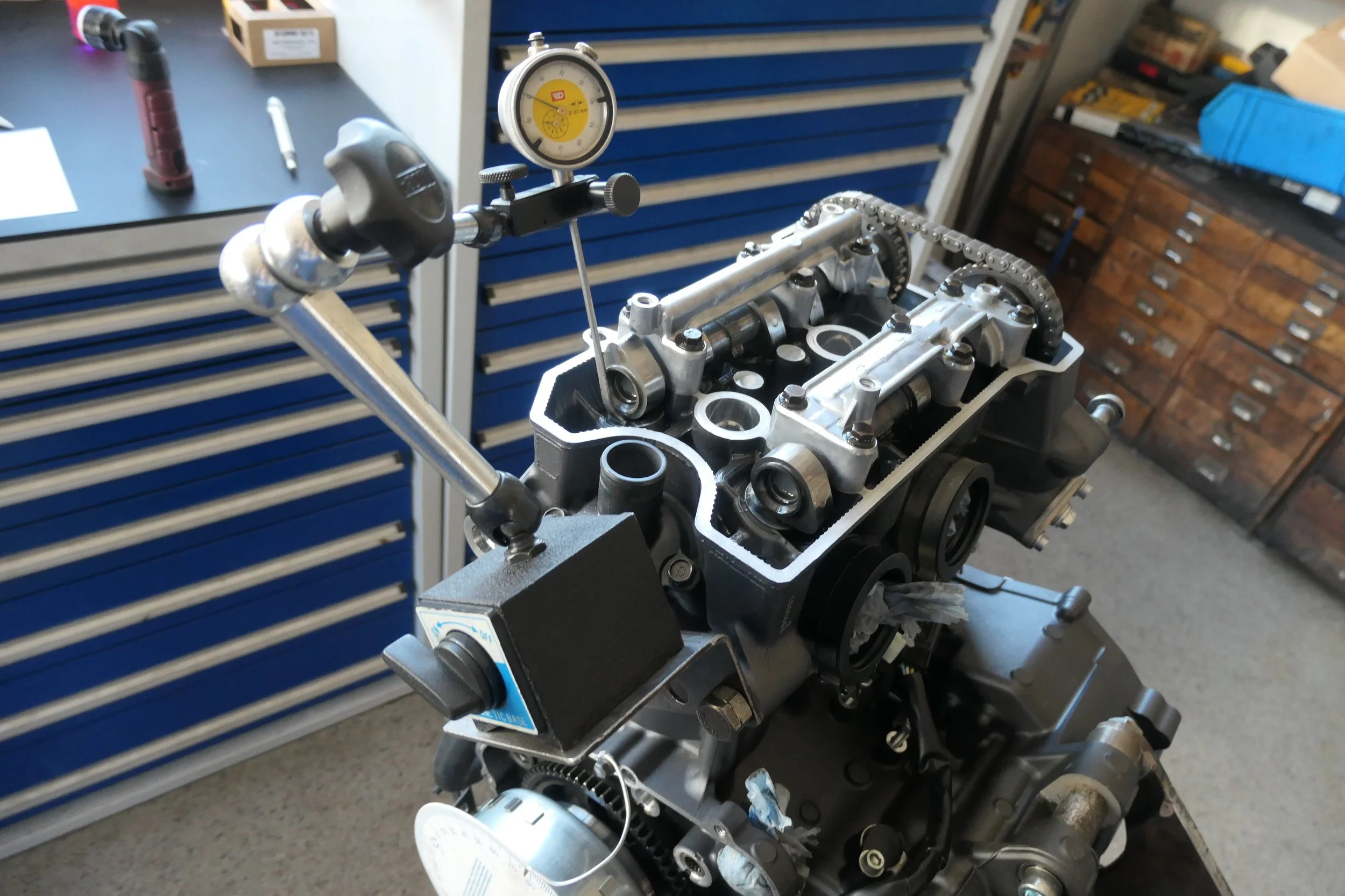 Benzinmotorblock auf Werkbank, mit Messgerät am Zylinderkopf, in Werkstatt mit blauen Schubladenschränken im Hintergrund.