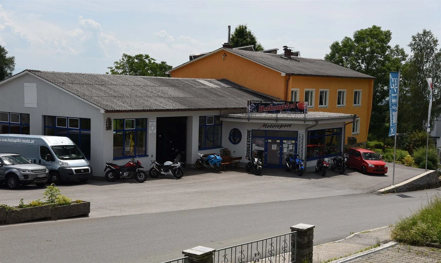 Motorsportgeschäft mit Motorrad- und Autoausrüstung, Parkplätzen und Flaggen, zweistöckiges Gebäude mit gelber und weißer Fassade, signiert mit 'Holzrapfel' und 'Bridgestone'