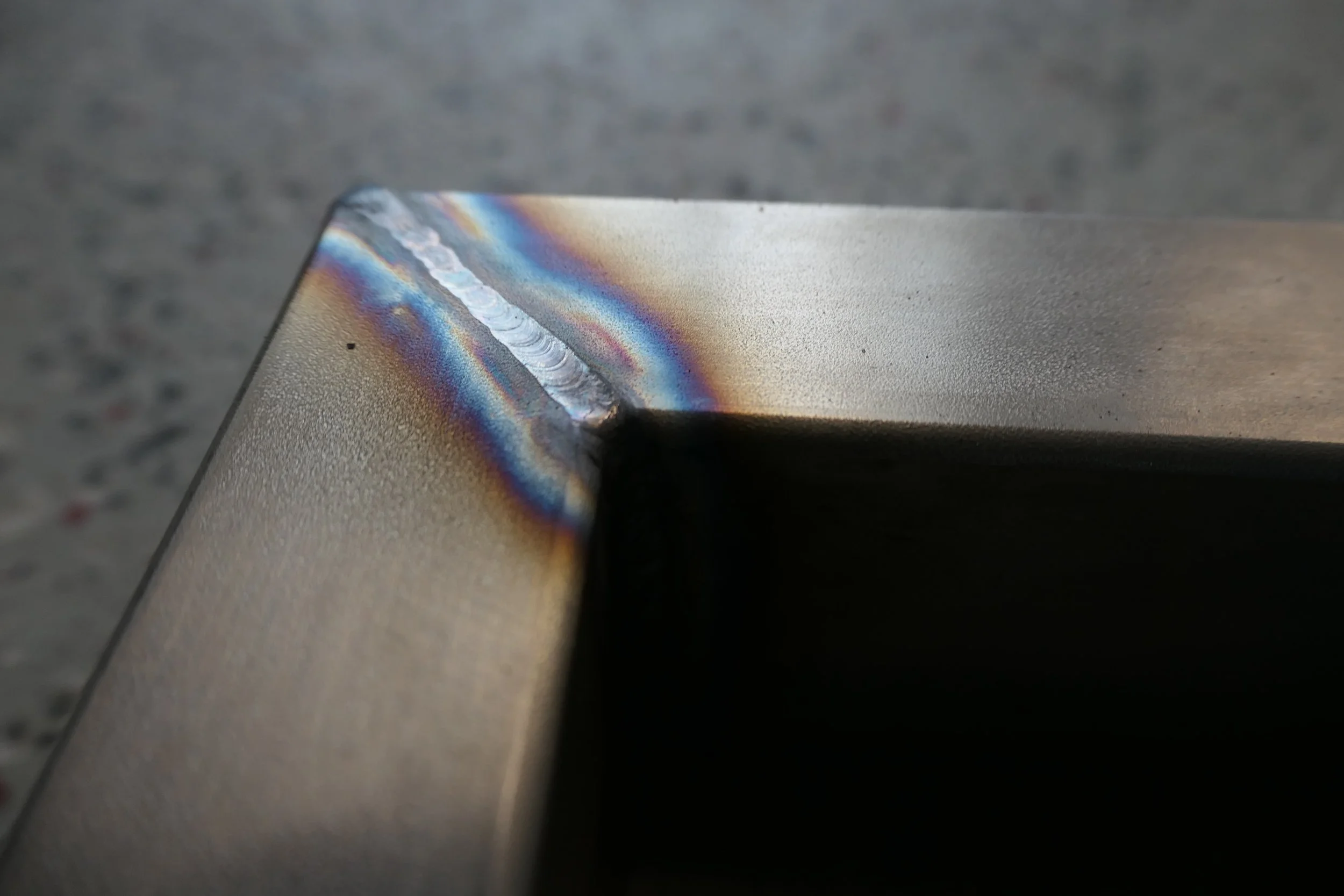 Nahaufnahme einer Stahlkante mit Schweißnaht, die bunte Wärmefarben aufweist, auf einem grauen, leicht geriffelten Hintergrund.