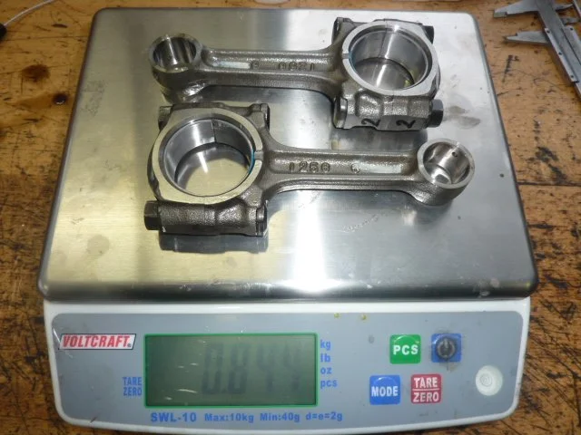 Ein metallisches Bauteil, wahrscheinlich eine Motorrad- oder Automobilkomponente, auf einer digitalen Waage mit Nullstellung, auf einem Holztisch.