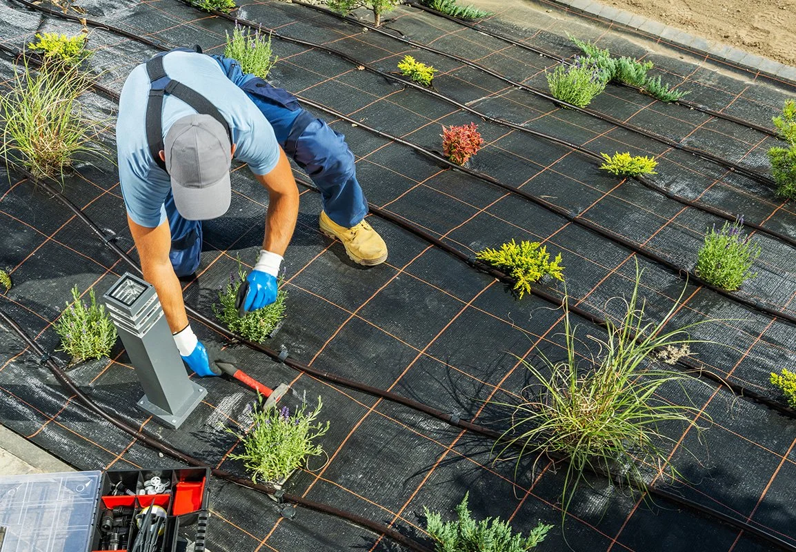 A person working on a garden bed, planting or tending to small plants on black weed fabric, with drip irrigation lines and a small solar-powered landscape light nearby.