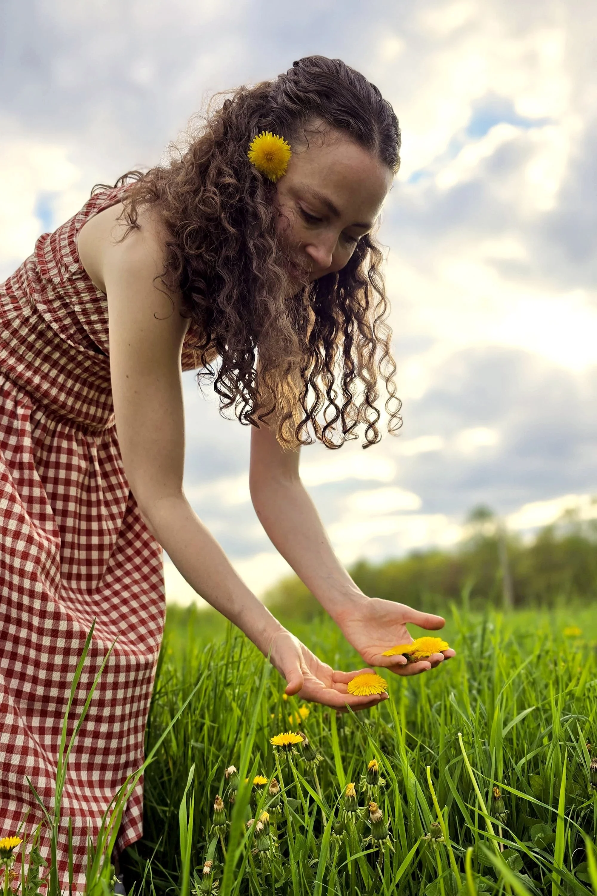 A woman with curly hair and a yellow flower in her hair bending down to pick yellow dandelions in a grassy field, with a cloudy sky in the background.