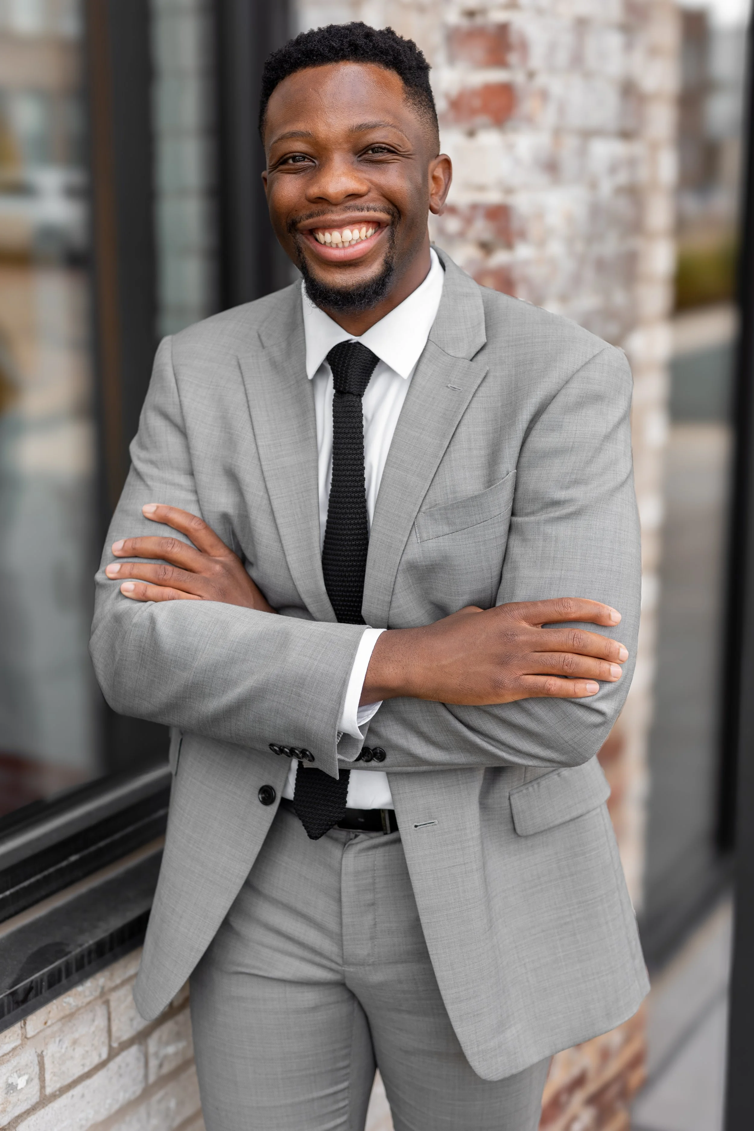 A smiling man in a gray suit with a white shirt and black tie standing with arms crossed in front of a brick and glass building.
