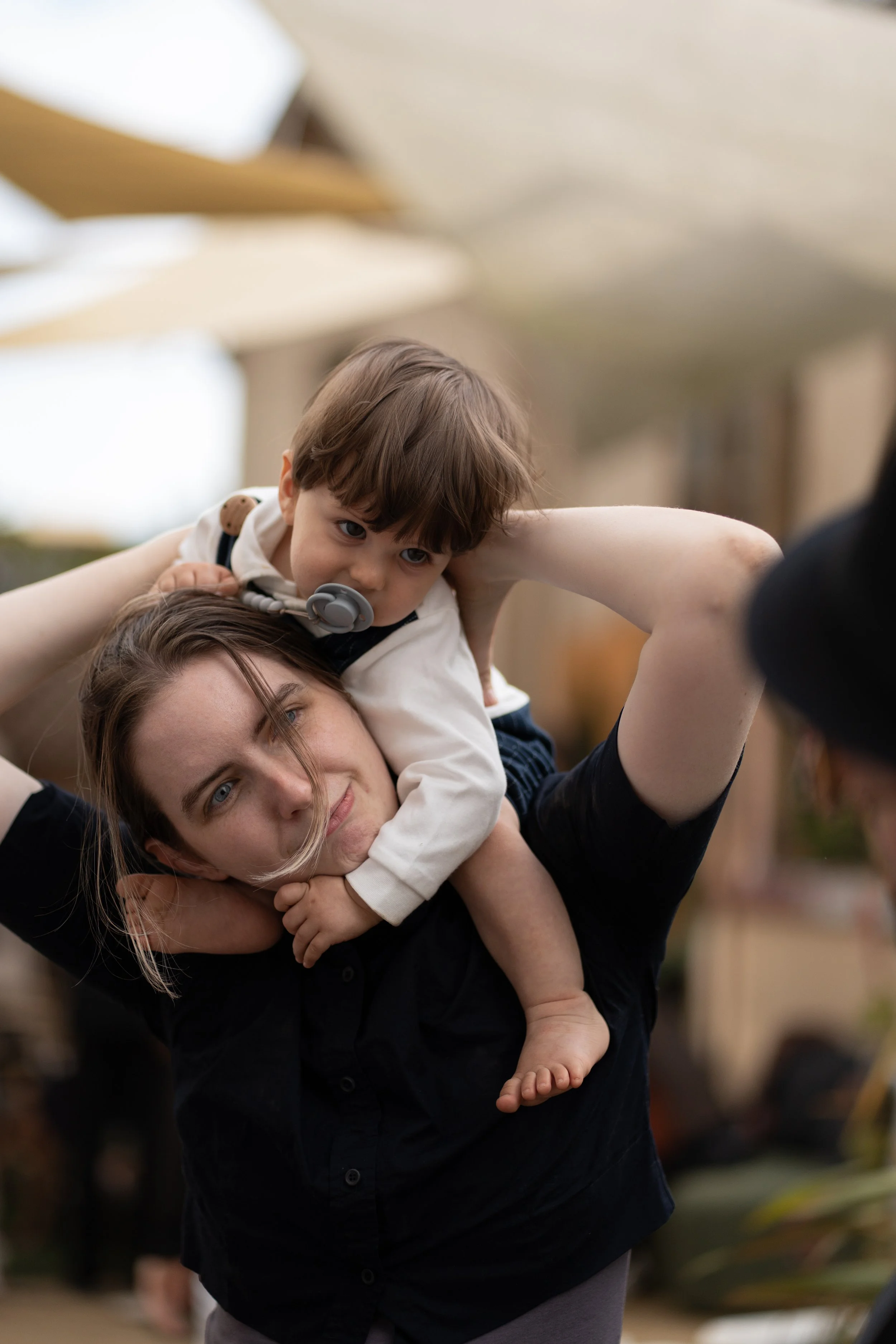 A woman is holding a young boy on her shoulders. The boy is leaning forward and resting his head on her head, with a pacifier in his mouth, and looking down. The woman is smiling while looking at the camera. The background is blurred.